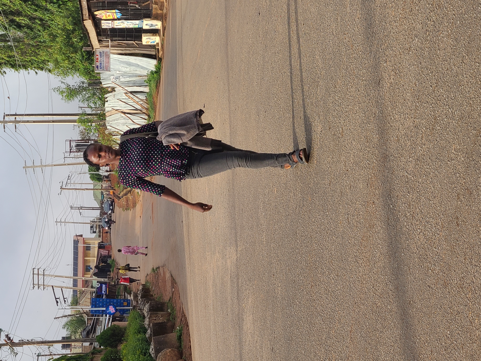 A medium-wide shot shows a person walking on a paved road in Jos, Nigeria. The person, who is the main subject, has dark skin and is wearing a dark, polka-dot shirt, gray pants, and sandals. A gray jacket is slung over their shoulder. The road appears to be a public street, stretching into the distance with other people walking in the same direction as the subject. Buildings, some with solar panels, line the left side of the street, while vegetation grows along the edge of the road on both sides. Utility poles with wires are visible against a slightly overcast sky. The location is determined to be Jos, Nigeria, through details like the local architecture, infrastructure, and the overall urban landscape.