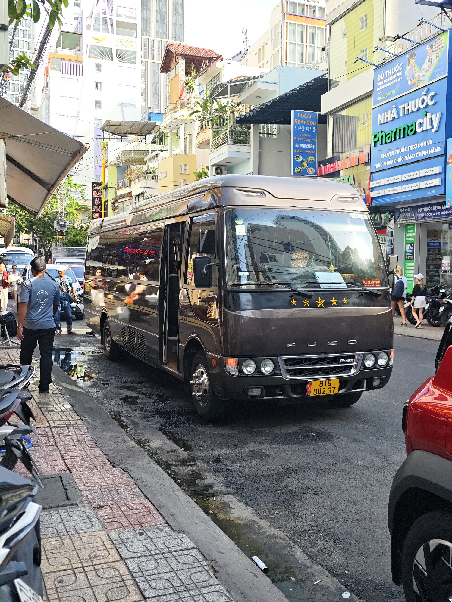 In Nha Trang Ward, Vietnam, a brown Fuso Rosa minibus is parked on the right side of a wet, asphalt street, facing forward. The minibus has its door open, and people are visible inside. To the left of the minibus, a man wearing a gray t-shirt with a white number "9" and black pants stands on a patterned tiled sidewalk, facing away from the camera. Several motorcycles are parked on the sidewalk behind him. A red car is parked to the right of the minibus. In the background, buildings of varying heights line the street, with signs and awnings visible. Overhead wires and branches of trees are also present. The sun is casting a bright glare on the scene.