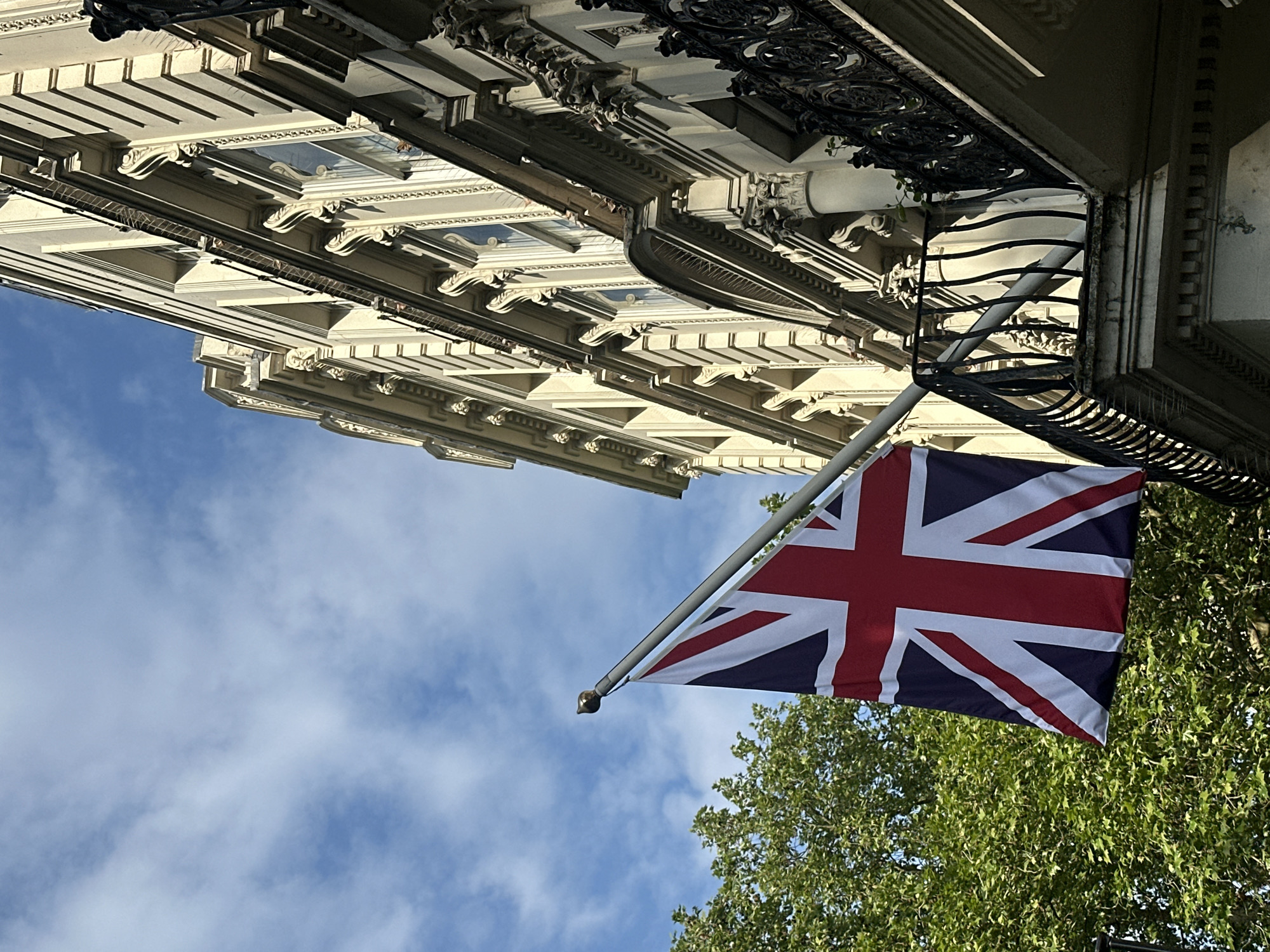 The image depicts a view looking upwards towards a building in London, United Kingdom. The building features ornate architectural details, including decorative moldings, windows, and a wrought-iron balcony. A Union Jack (British flag) hangs prominently from a pole attached to the building. The flag is partially obscured by the foliage of a tree. The background shows a blue sky with scattered clouds.