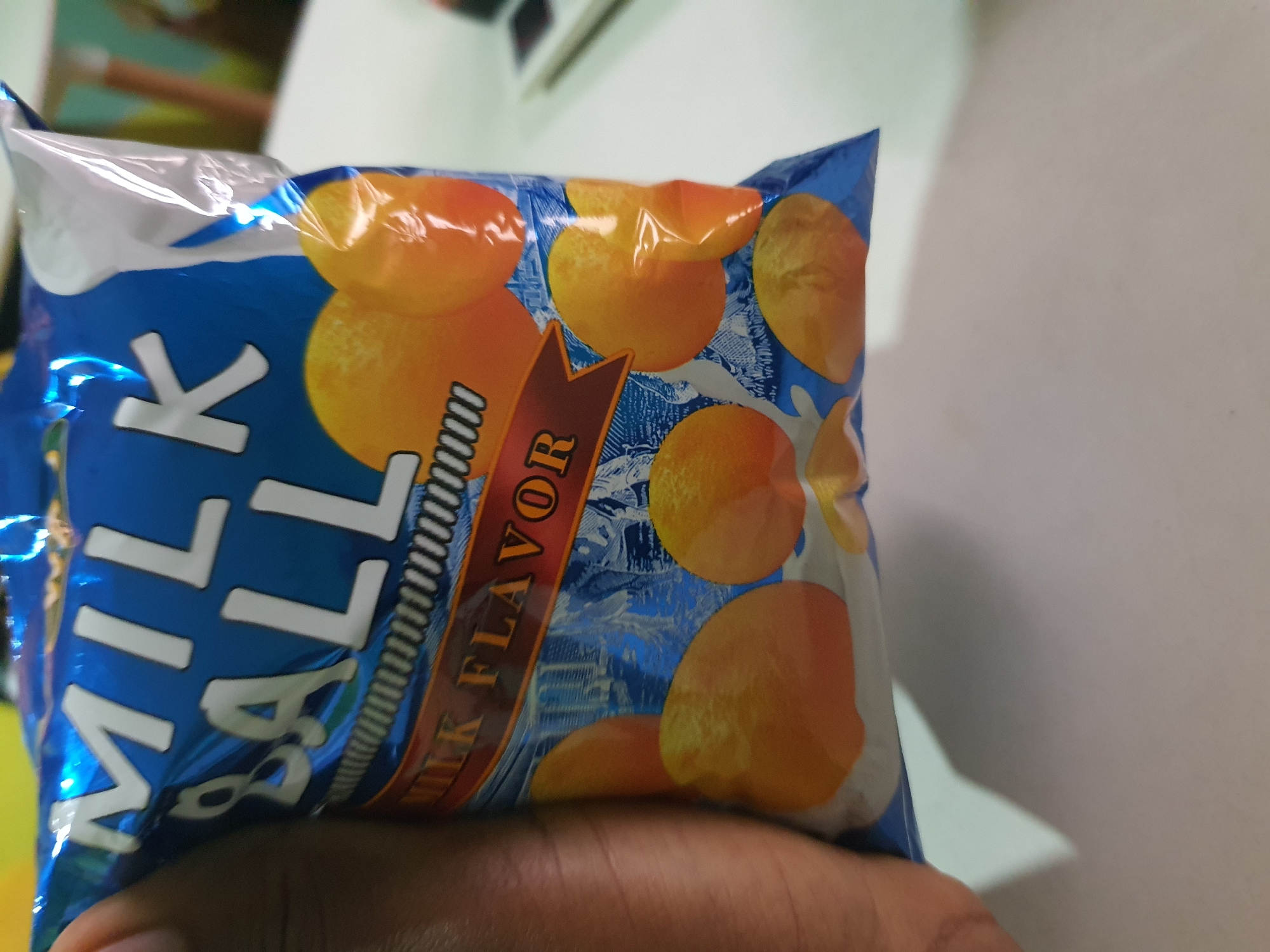 The image shows a close-up of a blue and white plastic bag with "MILK BALL" printed in large white letters. Below this, "MILK FLAVOR" is written in orange text on a red ribbon. The bag appears to be filled with small, round, orange-colored snacks, likely the milk balls. A person's hand, with dark skin, is holding the bag on the left side of the frame. The background is out of focus, but appears to be an indoor setting with a white surface, possibly a table. There is no identifiable information to place the image specifically in Palmgrove, Nigeria.