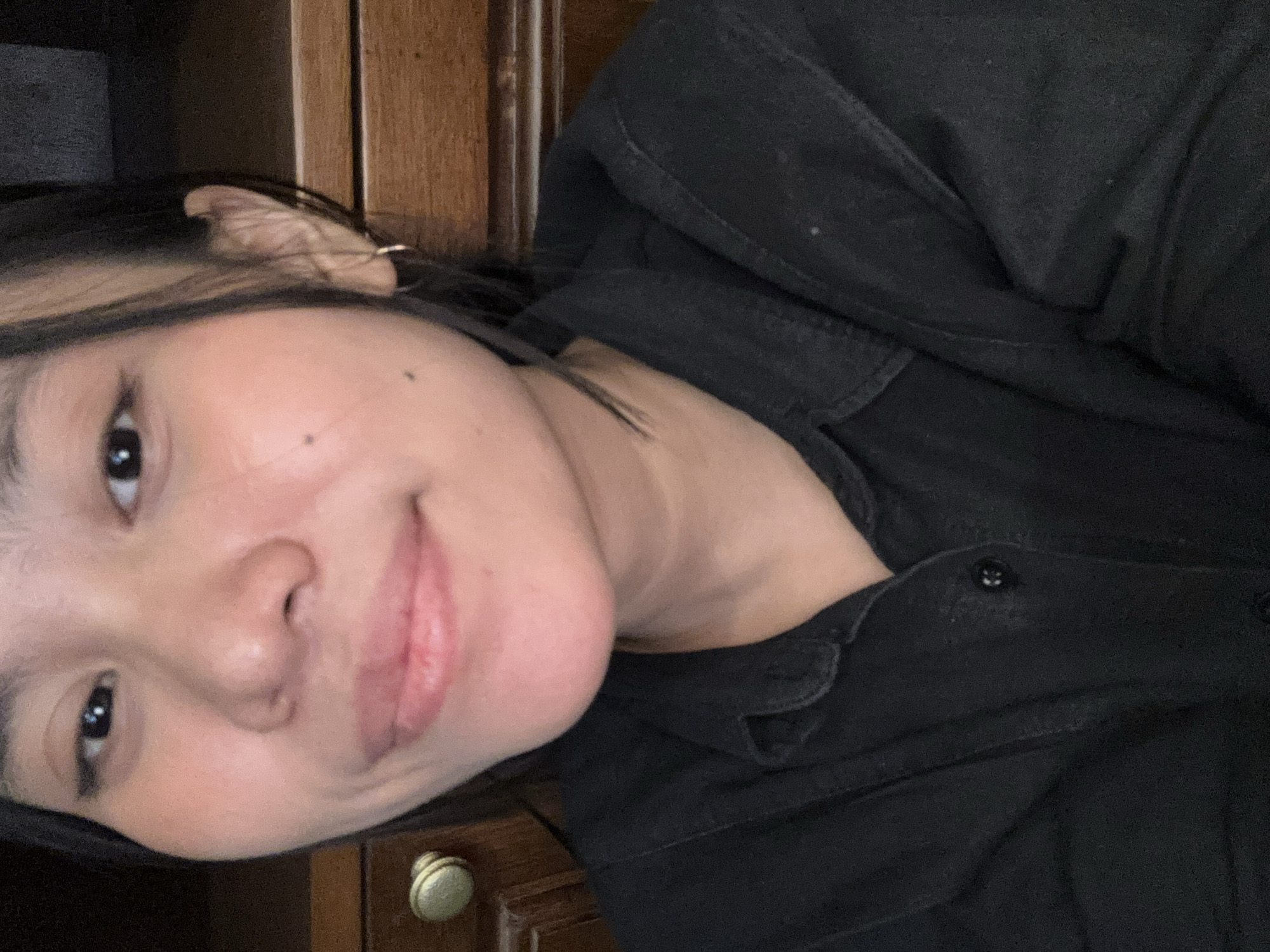 A close-up, head-and-shoulders selfie of a young woman with dark hair, fair skin, and light makeup is presented. She is wearing a black collared shirt. Her gaze is directed towards the camera with a slight smile. The background consists of a wooden cabinet with a brass knob on the left, and a dark, out-of-focus area on the right. The image appears to have been taken indoors.