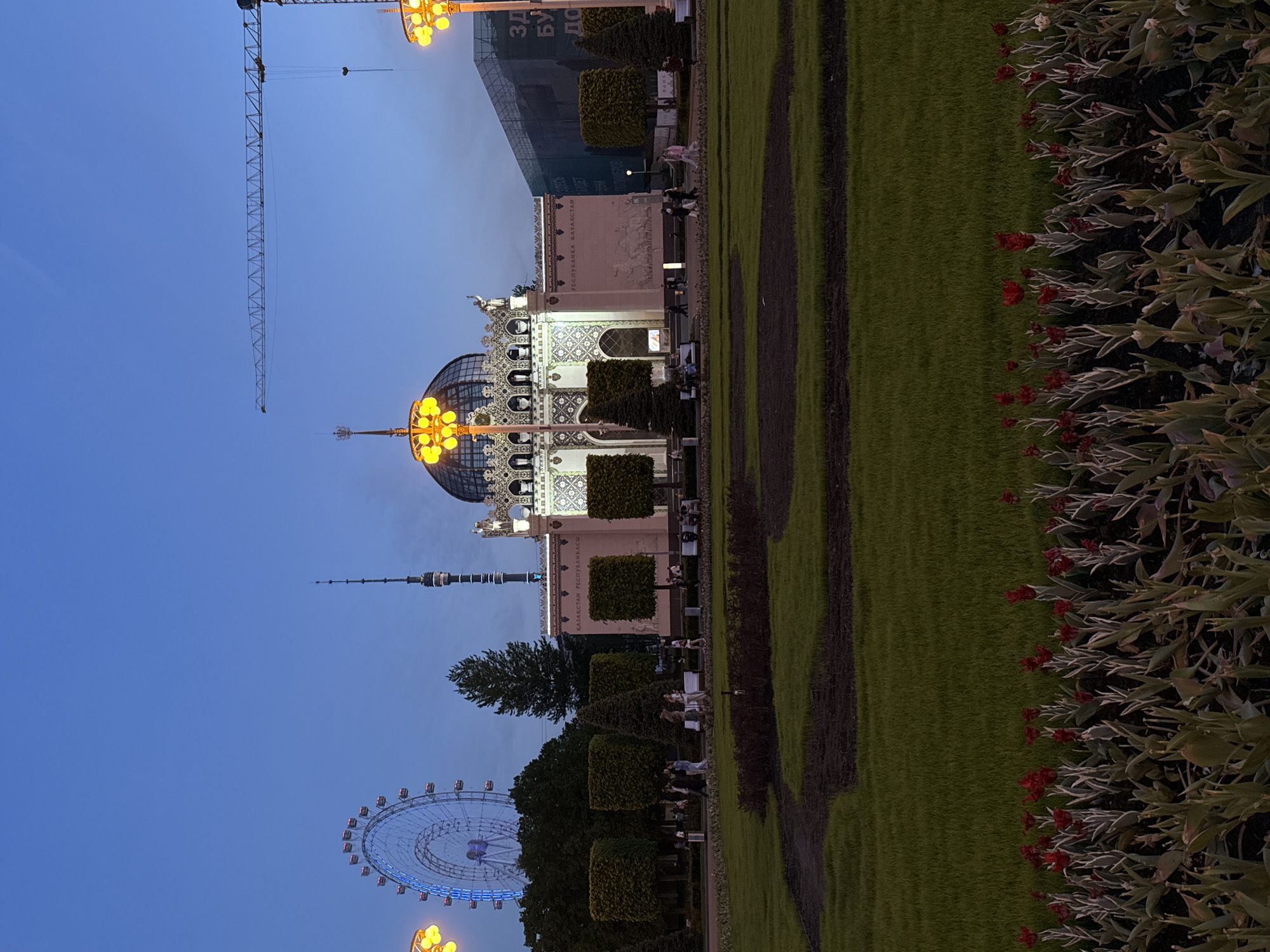 The image shows a park in Moscow, Russia, during the evening. In the foreground, there are flower beds with red flowers. Adjacent to the flowers is a well-manicured lawn. Further back, the Kazakhstan pavilion stands, illuminated with decorative lights. In the background, there is a Ferris wheel, the Ostankino Tower, and a construction crane under the twilight sky. People can be seen walking and sitting along the pathways in the park.