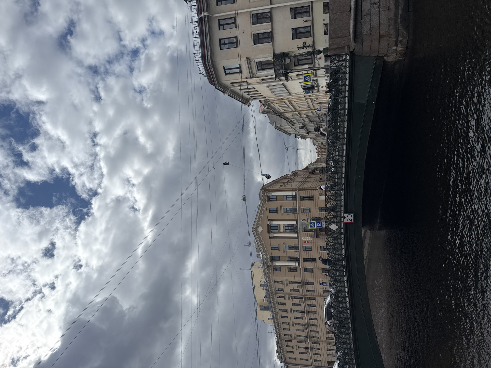 The image shows a bridge over a river in Saint Petersburg, Russia. The sky is cloudy with patches of blue. Buildings line the riverbanks, and power lines cross the sky. There are people standing on the bridge. A 2.0 height restriction sign is visible on the bridge railing. There are parking signs on the building. A pedestrian crossing sign is visible on the building.