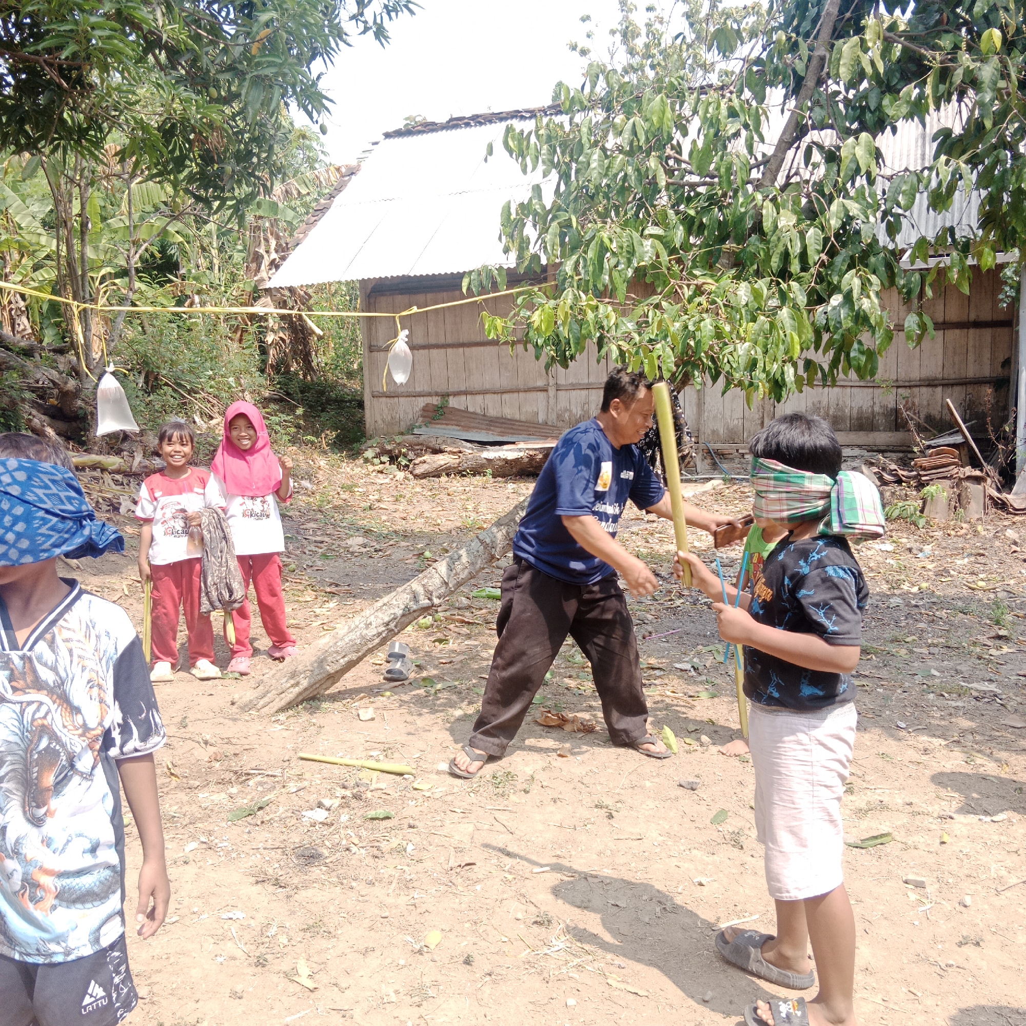 The image depicts an outdoor gathering in Tegowanu Kulon, Indonesia, likely a children's game or event. In the center, a man wearing a blue t-shirt and dark pants is holding a long wooden stick, seemingly guiding a child who is blindfolded with a checkered cloth. The child, wearing a black t-shirt and light-colored shorts, also holds a stick. To the left, two young girls are standing, one wearing a pink hijab and the other in a t-shirt and red pants, both smiling. Another child, partially visible on the far left, is also wearing a blue patterned blindfold. The environment is dusty and unpaved, with scattered leaves and debris on the ground. Several trees with lush green foliage are present, and a simple wooden building with a corrugated metal roof is visible in the background. A yellow rope is strung between two points, with a white plastic bag hanging from it. The overall scene suggests a rural or semi-rural setting in Indonesia.