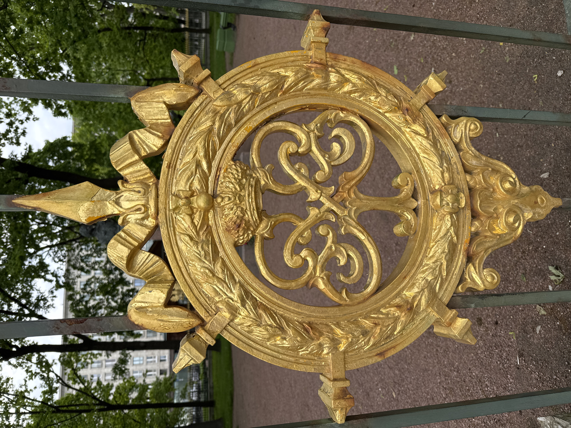 The image presents a close-up view of an ornate, gold-colored decorative medallion, likely part of a historic gate or fence in Saint Petersburg, Russia. The central feature is a circular design with intricate, intertwining scrollwork that vaguely resembles a stylized key or elaborate monogram. Within this central circle, a relief of a lion's head or similar mythological creature is visible on the left side.
Encircling the central motif is a broader ring adorned with a continuous laurel wreath pattern. Radiating outwards from this ring are various decorative elements, including pointed finials, gracefully flowing ribbon-like forms, and classical foliage motifs, all cast in the same striking gold material. The gold finish shows signs of aging and wear, with some areas of minor discoloration and chipping, suggesting its historical significance.
The golden ornament is attached to robust, dark green horizontal metal bars, which form part of a larger fence or gate visible in the foreground and background. Beyond the gate, a lush green park or garden environment is evident, filled with numerous trees bearing dense foliage. In the mid-distance, through the greenery, a modern-looking multi-story building with multiple windows is visible, contrasting with the classical aesthetic of the gate. The ground visible beneath the gate appears to be a gravel path. The weather is overcast, suggesting a cloudy daytime. No visible text is present in the image.