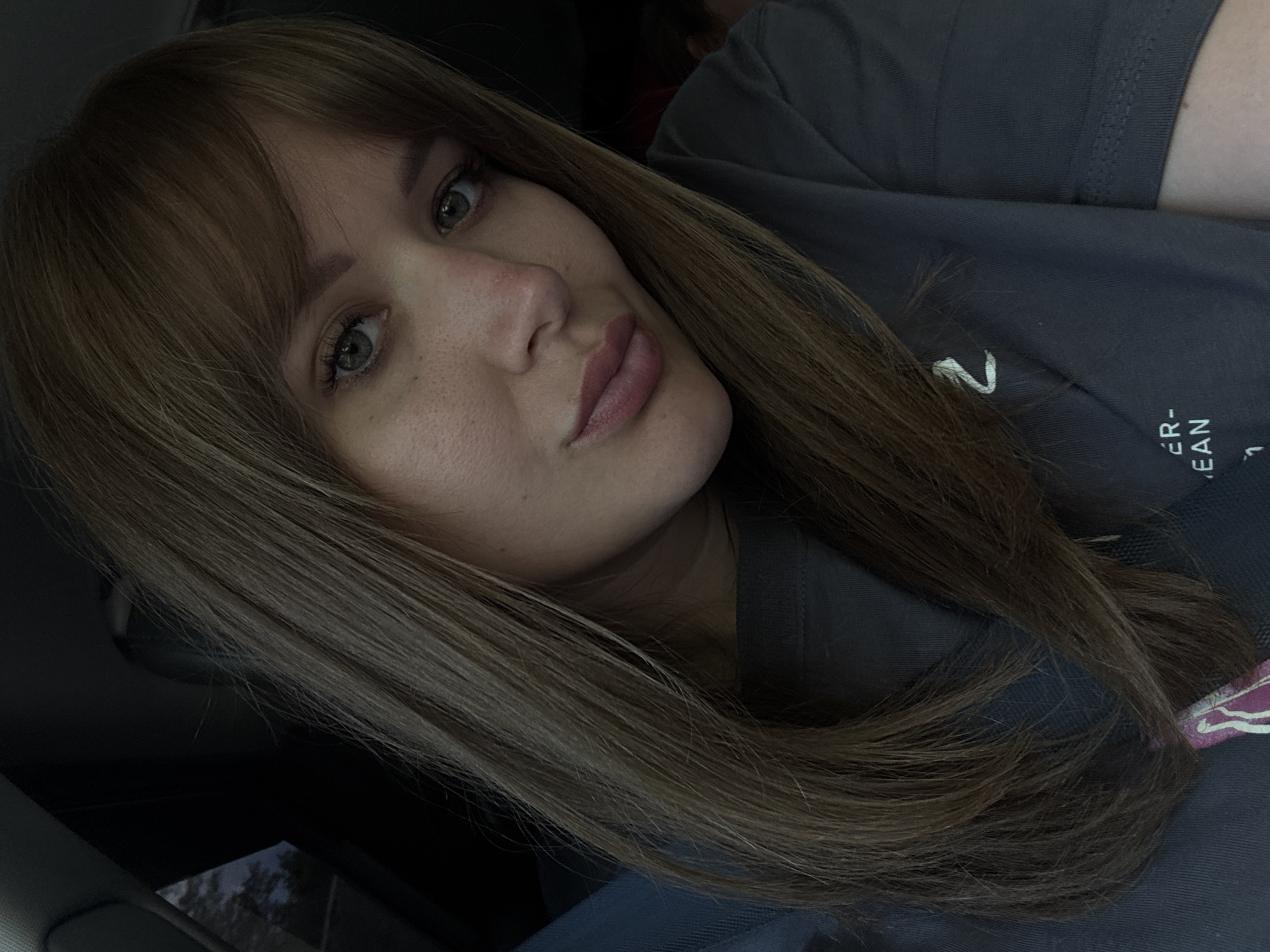 A close-up, slightly angled portrait of a woman with light brown, straight hair and bangs. She has fair skin, visible freckles on her nose, and light-colored eyes. She is wearing a gray t-shirt with a partially visible design on the front. The background is dark, suggesting an indoor setting, potentially a car interior with the edge of a window visible in the lower left. There is no evidence to suggest the location is Shaumyan, Russia. The image focuses on the woman's face and upper torso.