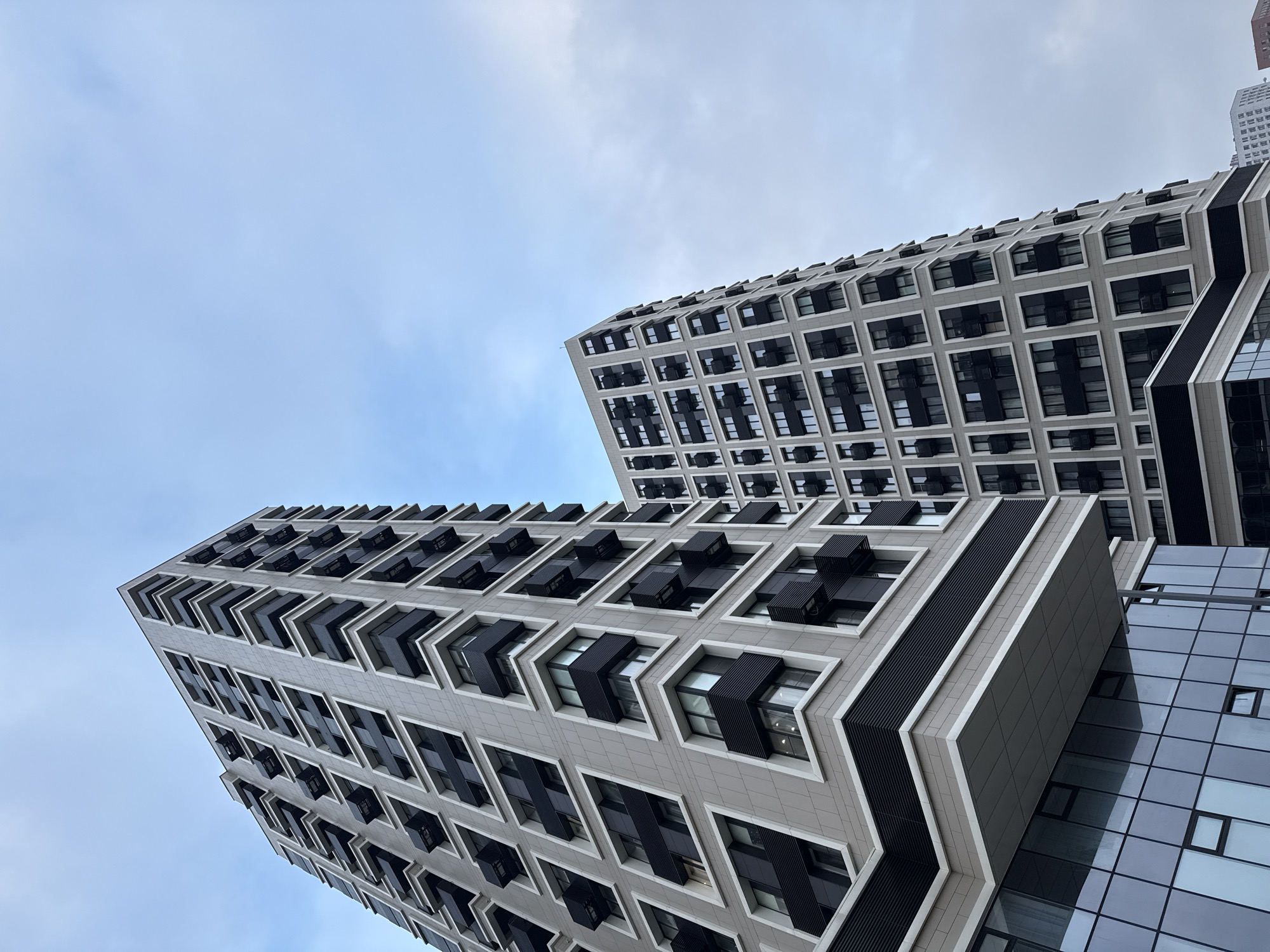 The image is a low-angle shot of two modern buildings against a cloudy sky. The buildings feature a grid-like facade with a pattern of rectangular windows and dark-colored protrusions, possibly balconies or architectural elements, extending from each window. One building has an additional glass section on its side. The buildings are located in Moscow, Russia.
