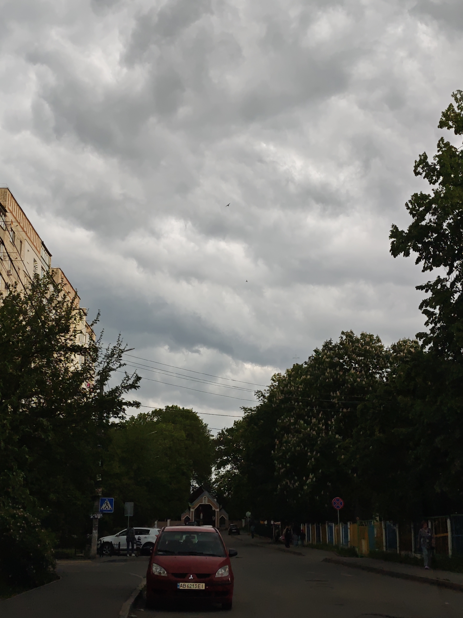 An eye-level, medium shot depicts a street in Vinnytsia, Ukraine, under an overcast sky. A red Mitsubishi Colt, license plate AB 6213 EI, is parked on the left side of the road, facing the viewer. A pedestrian crossing sign is visible on the left, near an intersection where a white car and a pedestrian are present. On the right, a colorful fence lines the sidewalk where several individuals are walking. Trees with green foliage frame the scene, obscuring parts of the buildings on both sides of the street. Overhead, power lines stretch across the road. A small church or chapel is positioned in the distance, centered within the street.