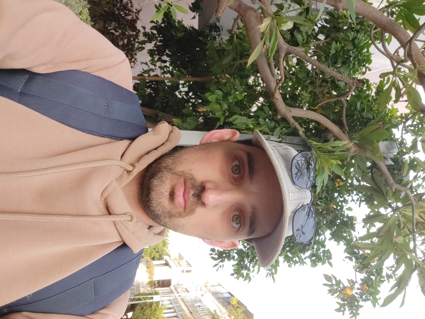 A selfie of a man wearing a tan hoodie with a blue backpack, a grey baseball cap, and sunglasses perched on the cap is angled upwards to include a tree with green leaves and small yellow fruit. In the background, buildings suggest an urban environment, consistent with the cityscape of Tel-Aviv, Israel.