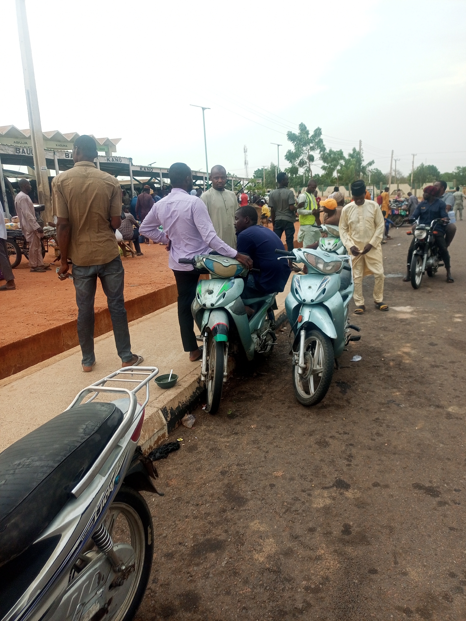 This image depicts a street scene in Gombe, Nigeria, near a marketplace or transportation hub. Several individuals are gathered in a group, some standing and conversing near parked motorcycles. The motorcycles are of the moped type, with two visible in the foreground that are light blue/silver in color. A third motorcycle can be partially seen in the foreground to the left.
The people in the scene wear a mix of traditional and casual clothing. Some are wearing traditional robes and hats, while others are in button-down shirts and trousers.
The environment includes an area with stalls that have signage in English indicating the destinations: Abuja, Kaduna, Bauchi, Kano, and Lagos. There is a concrete curb or sidewalk, and the street surface is paved but appears worn or damaged in places. The overall impression is a busy public space with a mixture of pedestrian and motorized traffic.
