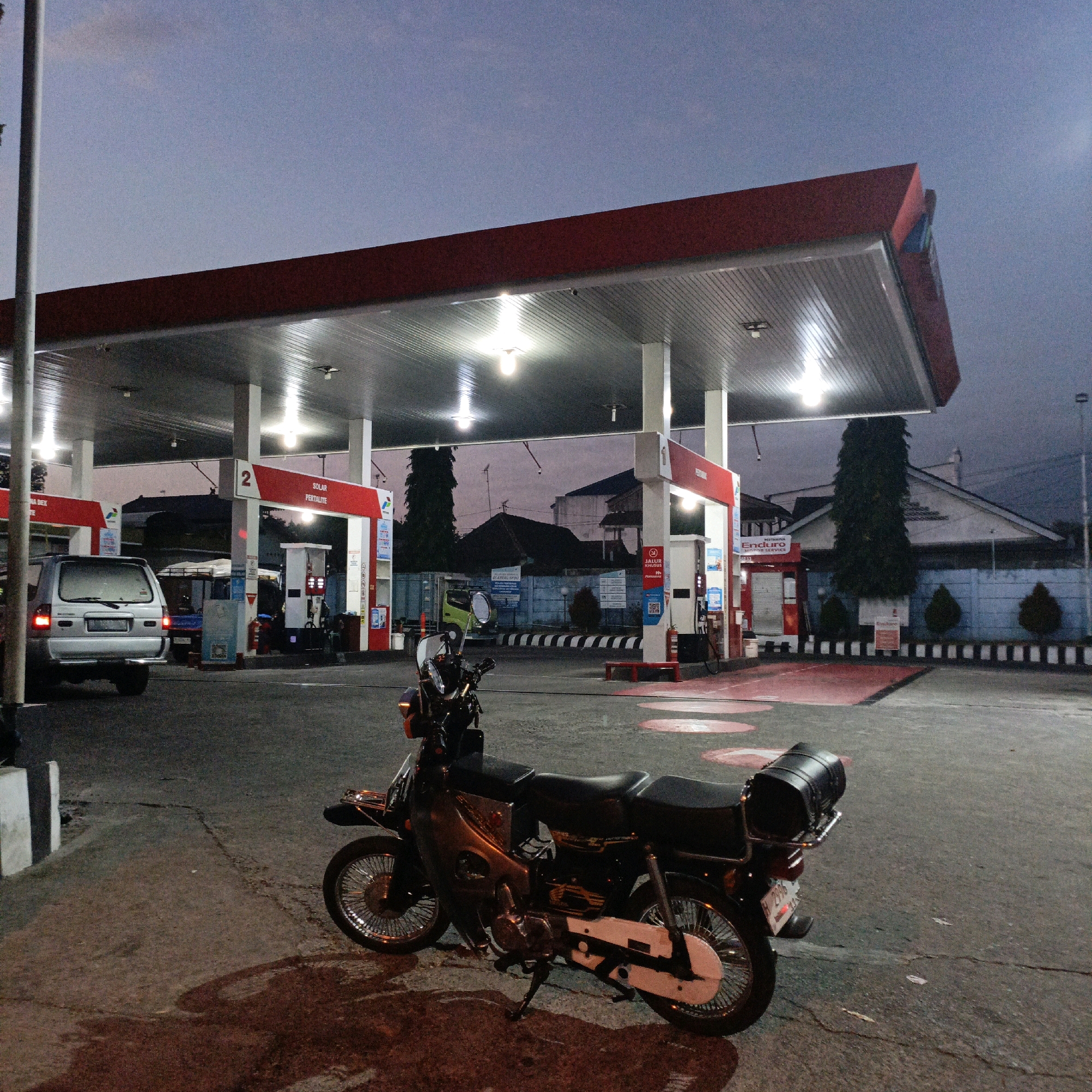 This image captures a gas station in Salatiga, Indonesia, during twilight. A black motorcycle is parked in the foreground, with its kickstand down and a black cylindrical storage box mounted on the rear rack. A silver SUV is visible in the midground, parked near a gas pump. The gas station features a red and white canopy with fluorescent lights illuminating the area. Red signage with the word "PERTAMINA" and the number "2" is visible on one of the support columns. In the background, buildings and trees are silhouetted against the dusky sky. The ground surface appears to be asphalt, with some cracks and scattered debris.