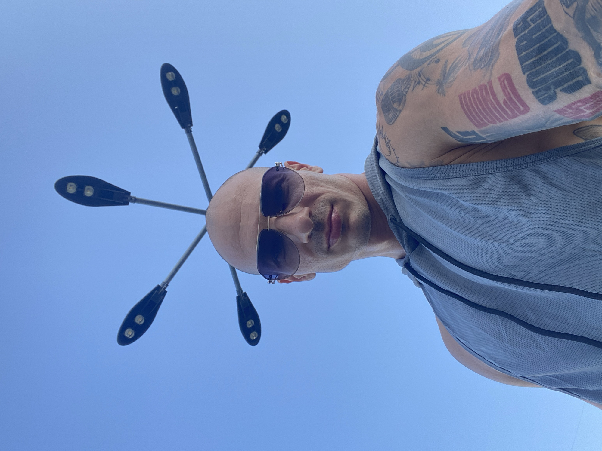 An outdoor, low-angle, close-up, waist-up shot shows a bald, white man wearing aviator sunglasses and a light blue, sleeveless hoodie. Six black, oval-shaped streetlights are positioned above his head, attached to thin metal poles extending from a central point. The man's left arm is visible, displaying multiple tattoos. The background consists of a clear, light blue sky. The image was taken in Pleven, Bulgaria.