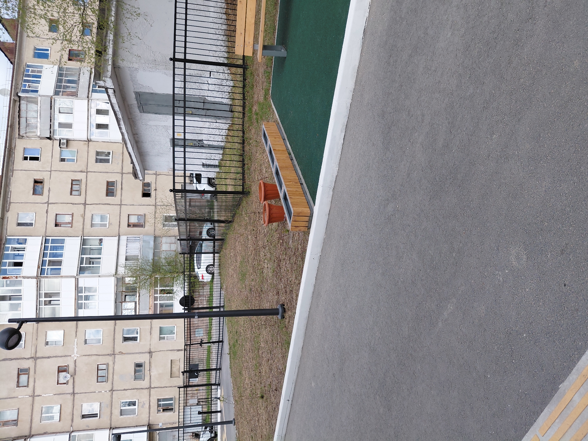 The image presents an angled, elevated view of an urban residential area in Nadym, Russia. The scene is dominated by a multi-story apartment building, characterized by its light-colored facade and numerous windows, many of which feature enclosed balconies with various glazing styles. An air conditioning unit is visible on one of the balconies.
In the foreground, a wide expanse of grey asphalt, likely a path or road, extends across the bottom and right side of the frame. It is clearly delineated by a white curb. Beyond the curb, a strip of dry, sparse grass leads to a black metal fence composed of vertical bars. Inside this fenced enclosure, a patch of bright green artificial turf is visible, on which a wooden bench with a grey metal frame is placed. Closer to the asphalt path, a long, rectangular wooden planter box holds two empty terracotta pots.
Further within the fenced area, a white car is partially visible, parked on a dirt or gravel surface. A prominent black street lamp post, topped with a round luminaire, stands near the fence. Sparse trees with light green foliage suggest a spring or early summer season, contrasting with the dry appearance of the grass. A small utility box or panel is affixed to a wall behind the fence.
The scene is static, devoid of people or specific activities. It appears to be daytime, with diffused lighting and no strong shadows. No visible text is present in the image. The architectural style and general environment are consistent with typical post-Soviet residential developments.