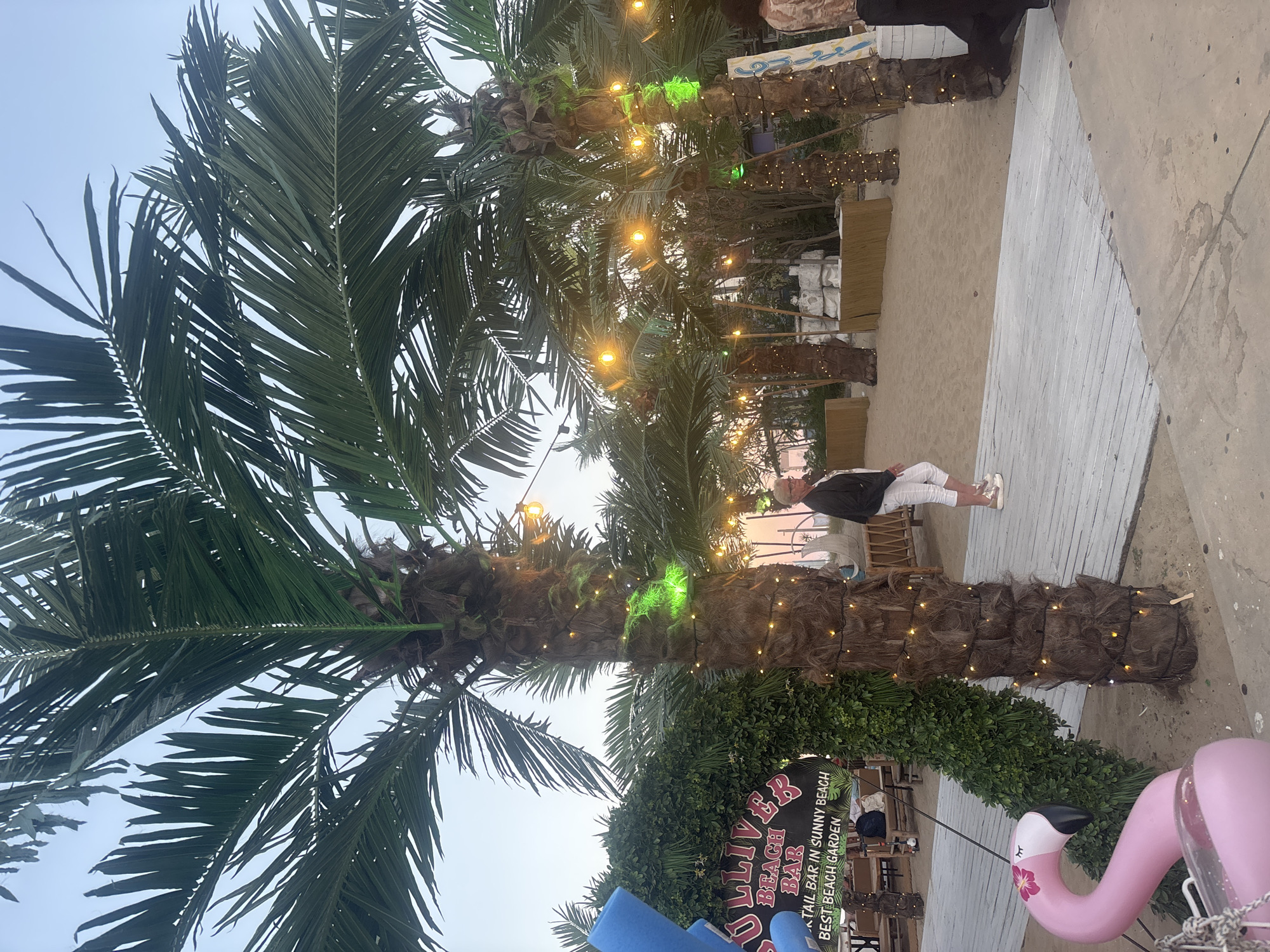 The image depicts an outdoor scene at Gulliver Beach Bar in Sunny Beach, Nesebar, Bulgaria. The setting is a sandy beach with several palm trees adorned with string lights. In the foreground, a pink inflatable flamingo is partially visible. A woman wearing white pants and a black jacket is walking on a wooden walkway. In the background, there are seating areas and structures suggesting a beach bar or venue. The sky is visible and appears to be twilight. The overall atmosphere suggests a relaxed, possibly evening setting.