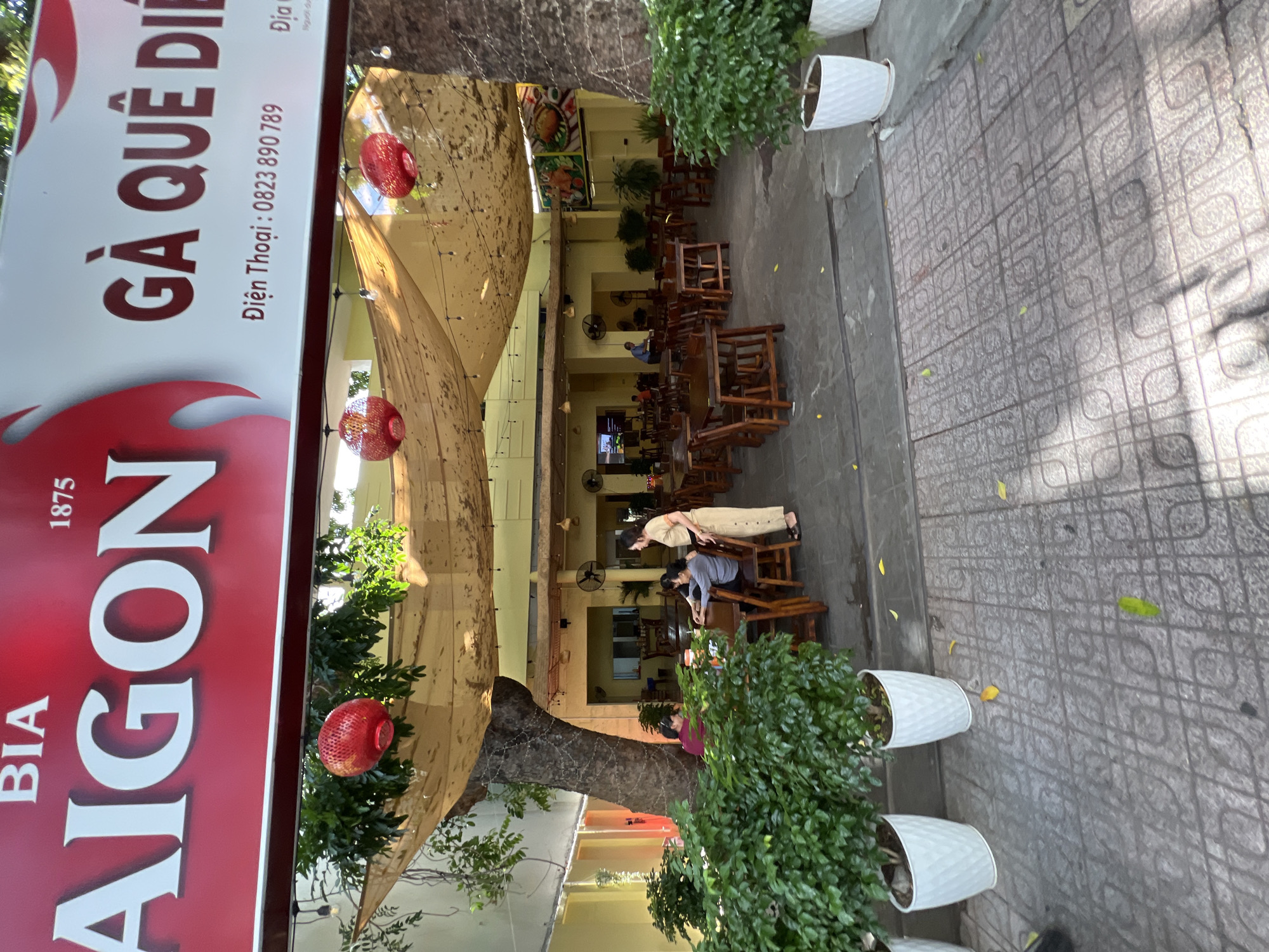 This is a medium shot, eye-level, exterior photograph of an outdoor seating area of a restaurant in Nha Trang, Vietnam, possibly named "Bia Saigon" and "Ga Que Dien". The restaurant features wooden tables and chairs arranged on a gray tiled patio under a large, beige fabric awning supported by posts and draped with string lights and decorative red lanterns. Green plants in white planters are placed at the front of the patio. Several patrons are seated at tables, and one woman in a beige dress is standing and looking towards the tables. The background shows a building with yellow walls and open doorways, containing more tables, chairs, and ceiling fans. A large tree with a thick trunk is situated on the left side of the patio, partially obscured by foliage and the awning. The overall atmosphere is casual and inviting, suggesting a sunny day.