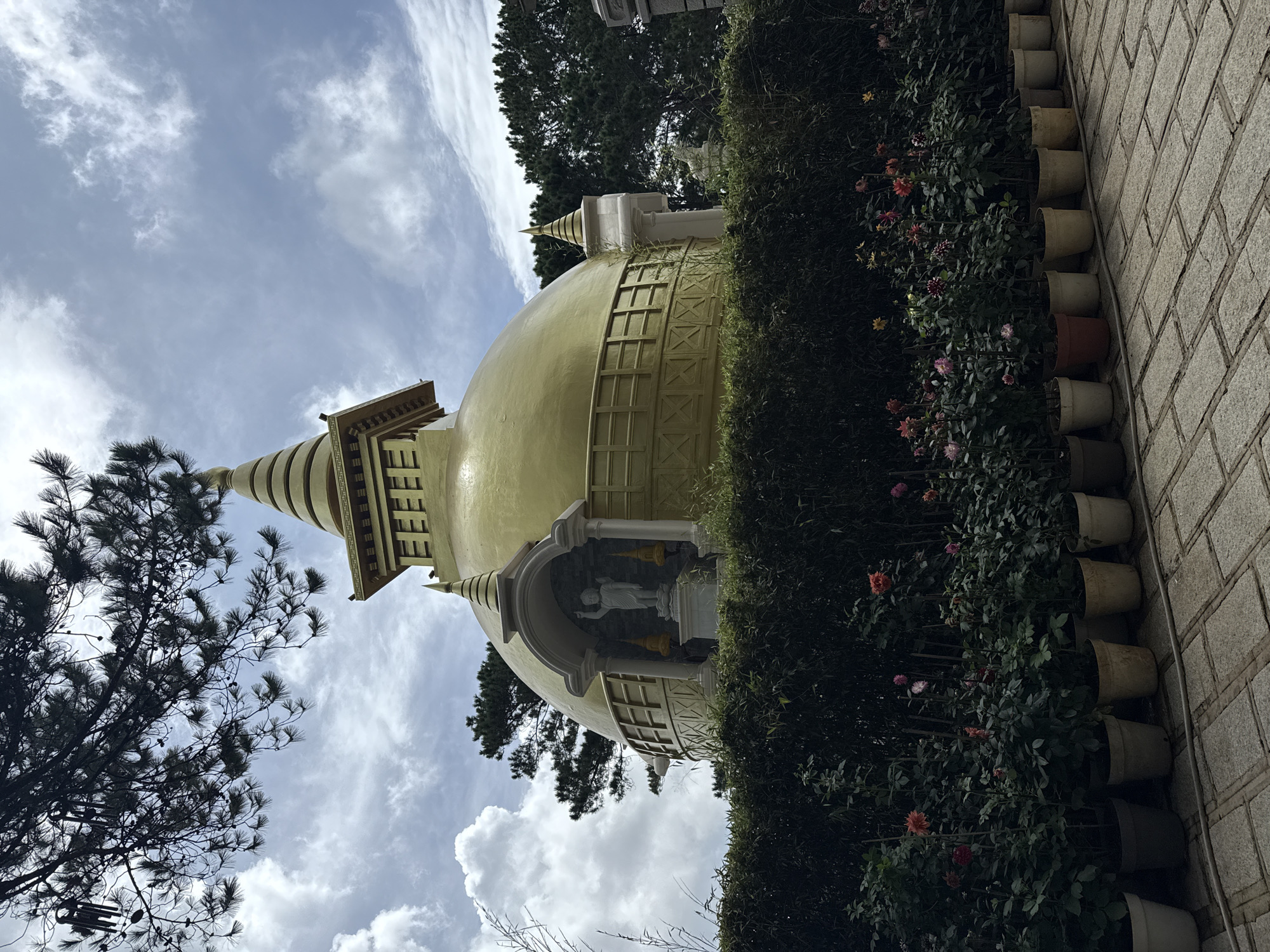 The image depicts a large, golden stupa with a tiered conical roof, set against a cloudy sky. The stupa is adorned with architectural details and features a niche containing a white statue of a standing figure, possibly Buddha, with two smaller conical finials on either side. Surrounding the stupa is a dense hedge of green foliage, and in front of the hedge, a row of potted plants with red, pink, and yellow flowers are arranged along a paved walkway made of stone tiles. Pine trees are visible in the background, with branches extending into the upper left corner of the frame. This stupa is identified as the Linh Phuoc Pagoda, a Buddhist temple located in Da Lat, Vietnam, renowned for its intricate mosaic work and unique architecture. There are no people visible in the image.