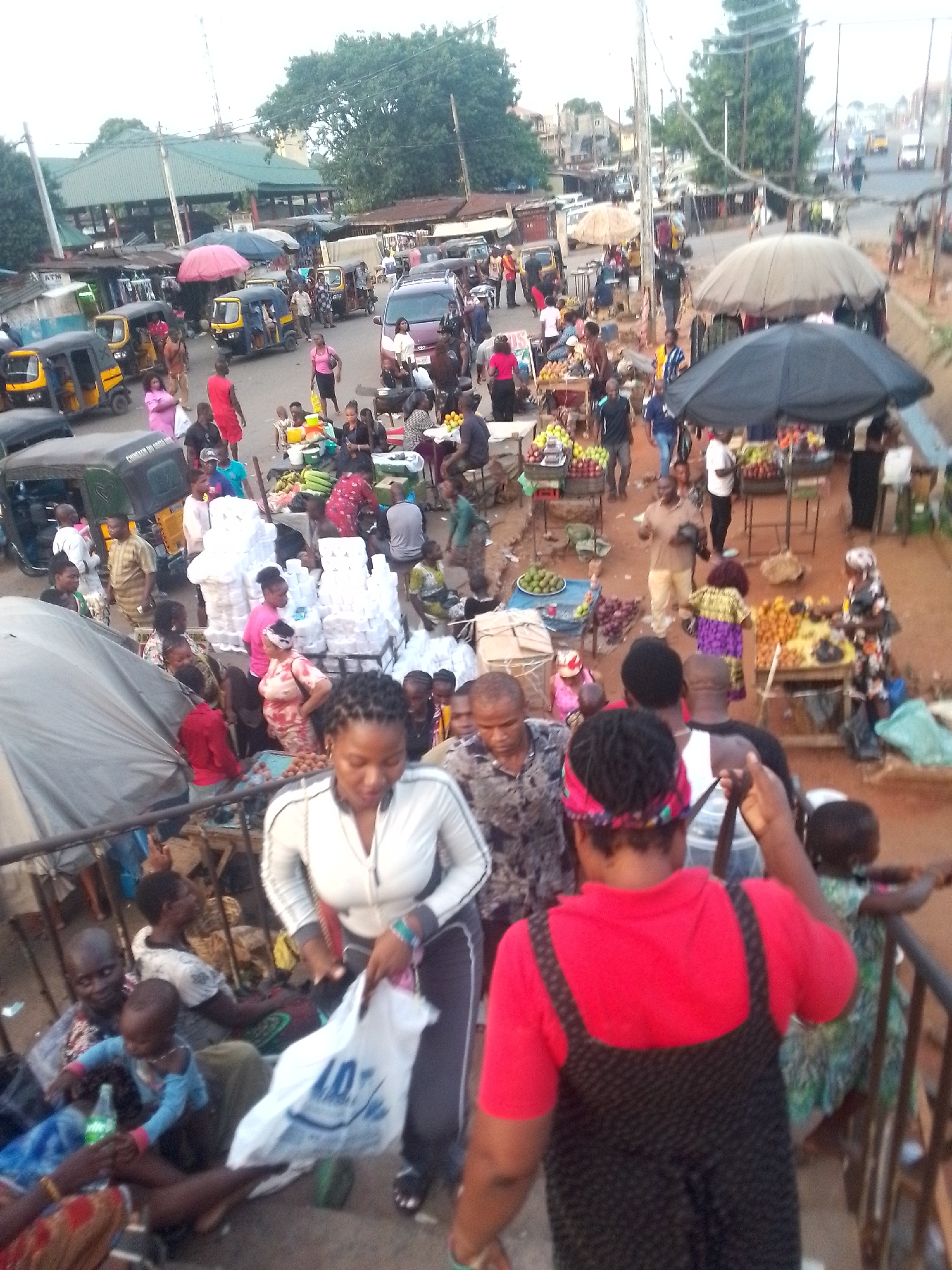 A bustling street scene in Unknown, Unknown shows a crowded marketplace from an elevated perspective. The ground is dusty, packed with people and vendors, several under large umbrellas. Market stalls display fruits and other goods. Vehicles including auto rickshaws navigate the crowded street. People are walking, selling, and interacting in the market. Buildings and trees are visible in the background.