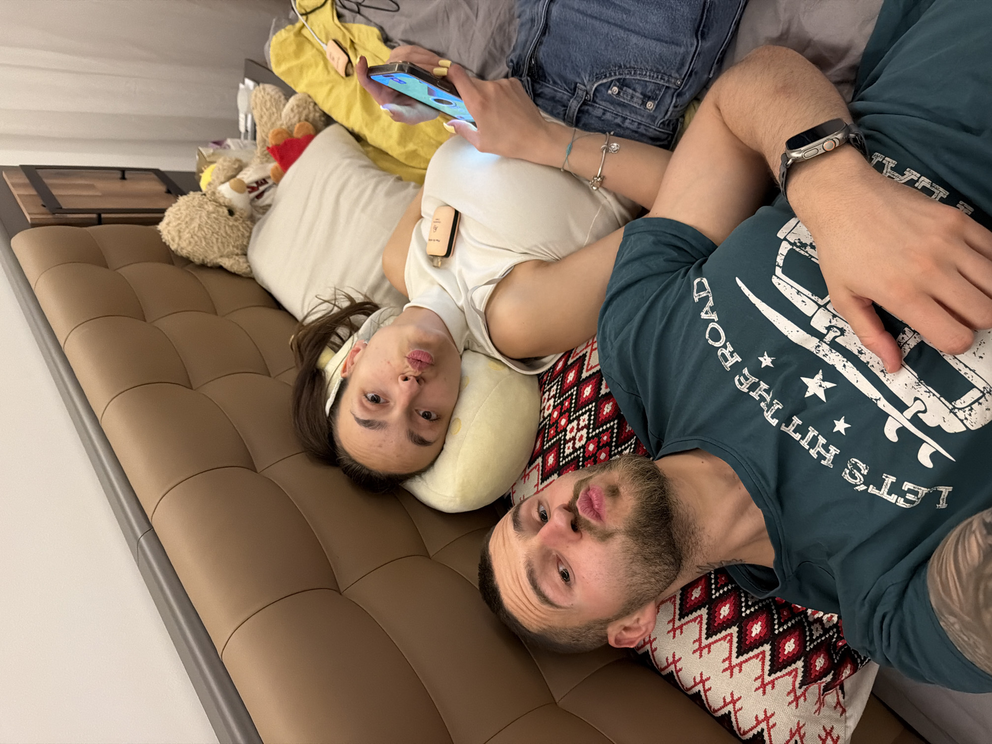 This media file, captured in Sofia, Bulgaria, shows two individuals, a man and a woman, relaxing indoors, possibly on a sofa or bed. The image is rotated 90 degrees counter-clockwise, making the subjects appear to be lying on their sides, looking towards the camera with playful, pouty expressions.
The woman is positioned in the upper part of the frame, wearing a white sleeveless top and blue denim jeans. She has long dark hair, partly held back by a light-colored headband, and wears a silver bracelet on her wrist. Her fingernails are painted yellow. She is actively engaged with a smartphone, holding it with both hands, its screen glowing brightly. A small, light-colored rectangular device, possibly an e-cigarette or portable charger with text "Pure Go" partially visible, is tucked into her top. She rests her head on a soft, light yellow/cream pillow. Behind her, several stuffed animals, including a teddy bear, are visible on what appears to be a bed or sofa backrest.
The man is in the lower part of the frame, with a beard and short dark hair. He is wearing a dark green long-sleeved t-shirt that displays white text and graphics. The visible text on his shirt reads "LET'S HIT THE ROAD" along with star motifs and an emblem that might be a surf or vehicle outline. A smartwatch with a dark band is worn on his left wrist, and a tattoo is visible on his right arm. He rests his head on a patterned pillow featuring red, black, and white geometric designs.
The furniture beneath them is a brown, tufted, leather-like surface, indicating a comfortable seating or sleeping arrangement. A simple wooden and metal side table or shelf is partially visible in the background. The overall atmosphere conveyed is relaxed and casual, with the individuals posing humorously for the camera.