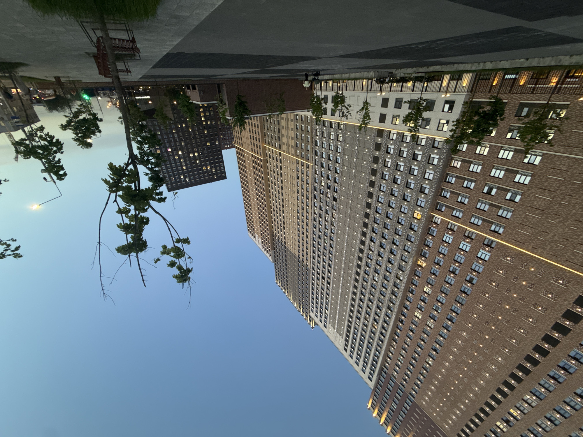 The image depicts a street scene in Moscow, Russia, likely taken in the evening or early morning, given the twilight sky. Two modern high-rise apartment buildings are visible, constructed with brick and stone facades, featuring numerous windows. The buildings display architectural lighting, enhancing the view. Trees line the street and a pedestrian walkway paved with grey and black tiles is in the foreground. A bench is visible in the upper-left corner. A street lamp is also present near the trees. No people are immediately visible.