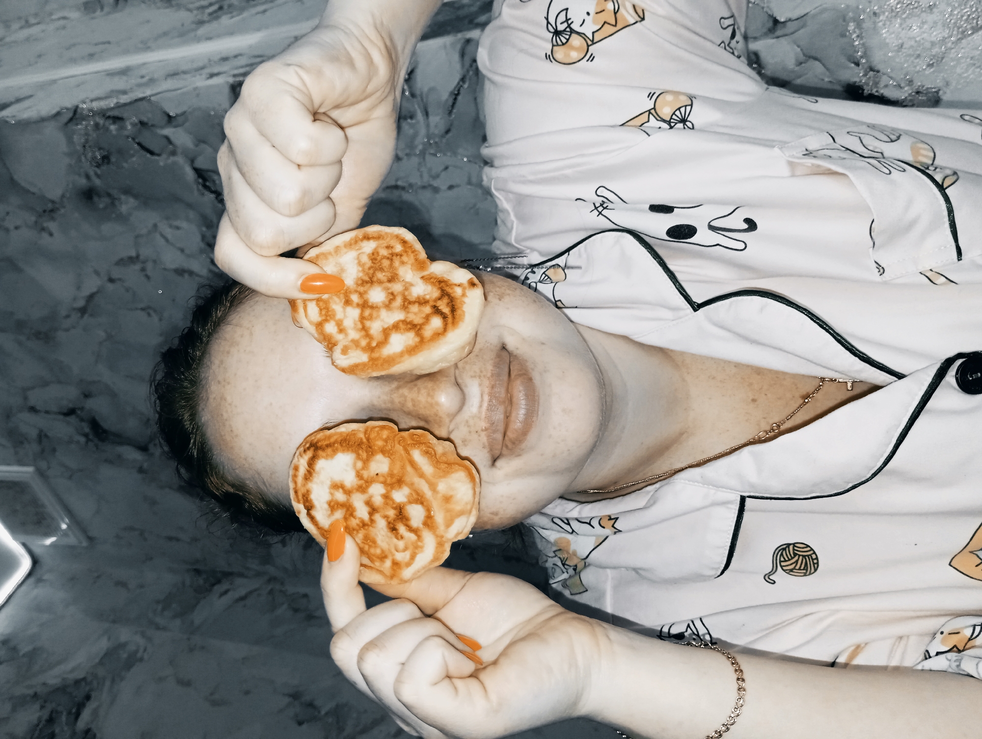 A woman is lying on her back in a bathroom with marbled walls and floor. She is wearing white pajamas patterned with cartoon drawings. She is holding a pancake over each eye. The woman has fair skin and freckles. She wears orange nail polish. A gold necklace and bracelet are visible. The location is presumed to be Yurgamysh, Russia.