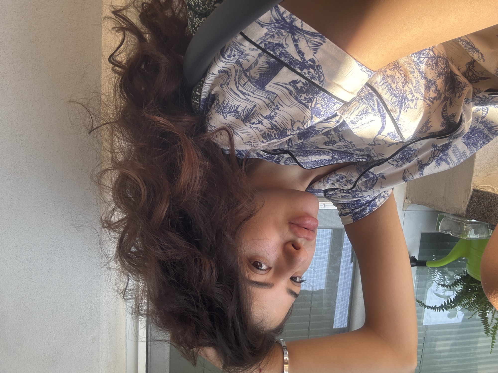 In a bright indoor setting, a young woman with long, wavy brown hair is captured in a relaxed pose. She is wearing a light-colored pajama top with a blue and white intricate pattern, possibly depicting a landscape or toile design. Her left arm is raised, with her hand resting on her head, and her gaze is directed towards the camera. The sunlight casts a warm glow on her skin, highlighting her features. Visible in the background is a window with blinds, a green watering can, and a small potted plant, suggesting a domestic environment. The overall scene conveys a sense of casual comfort. The image does not provide any specific clues that would allow for its location to be identified within the city of Plovdiv, Bulgaria.