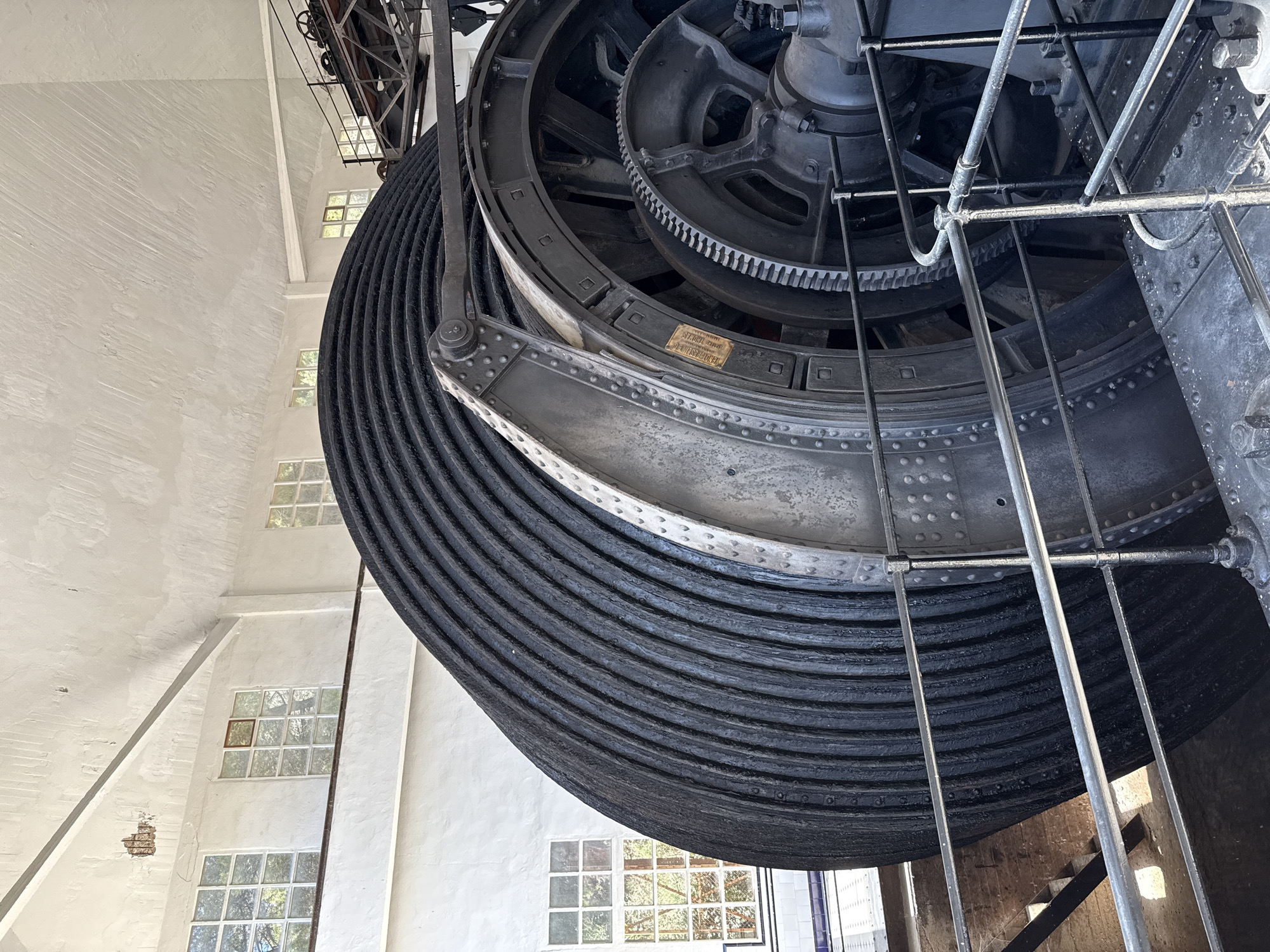 The image depicts the interior of the Salt Mountain Cultural Park in Cardona, Spain, specifically focusing on the large winding drum of an old mining machine. The drum is massive and covered in thick, black coiled rope, dominating the foreground and right side of the frame. It is held in place by heavy metal supports and a large gear mechanism is visible to its right, suggesting its operational purpose.

In the background, a white, industrial-style building with a high, textured ceiling and white-painted walls is visible. Several large, multi-paned windows with white frames are set into the walls. Through the windows, greenery and trees are discernible, indicating the presence of an outdoor environment beyond the structure. The overall impression is one of historical industrial machinery within a preserved mining heritage site. No people are present in the image.