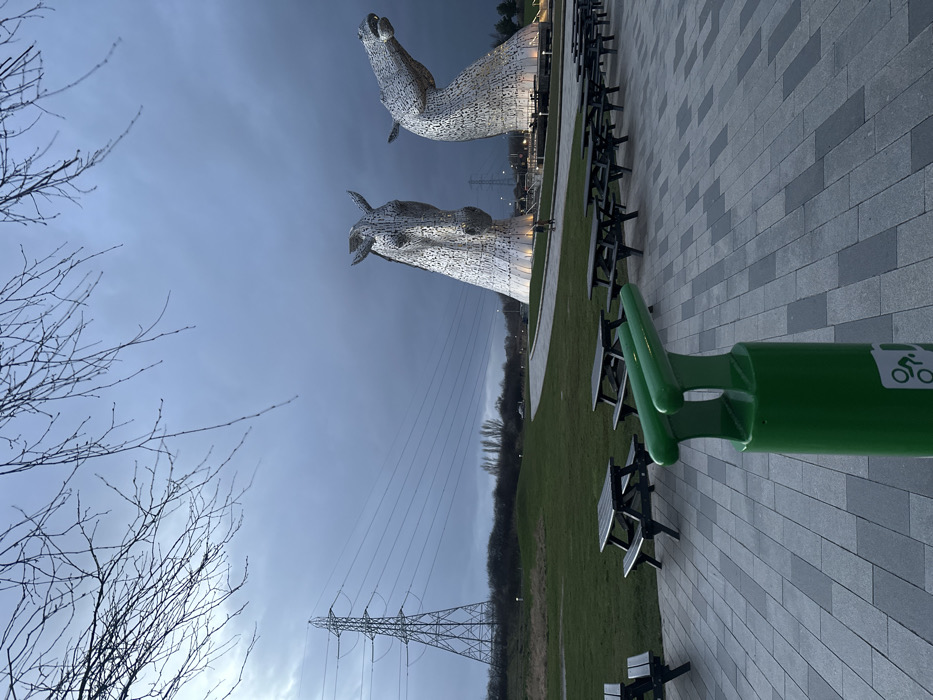 This image depicts The Kelpies, two colossal metallic horse head sculptures, situated in The Helix park in Falkirk, United Kingdom. The sky is dark grey and overcast, with bare tree branches visible in the upper portion of the frame. In the immediate foreground, a bright green cylindrical bicycle repair stand, featuring a white bicycle icon near its base and two upper protrusions, is centrally positioned.
Behind this stand, a series of dark wooden picnic tables with attached benches are arranged on a paved area composed of alternating light and dark grey rectangular slabs. Beyond the paved area, a manicured green grass lawn extends, with a paved pathway curving away to the right. A single, distant human figure, dressed in dark clothing, is walking on this pathway between the picnic tables and the sculptures.
The two Kelpie sculptures are the dominant subjects in the mid-ground, illuminated from their bases. The left sculpture presents a frontal view of a horse's head, while the right sculpture's head is angled upwards. To the far left, a large metal electricity pylon supports multiple power lines that stretch horizontally across the landscape. The distant horizon reveals scattered trees and shrubs. The lighting suggests late afternoon or an overcast day.