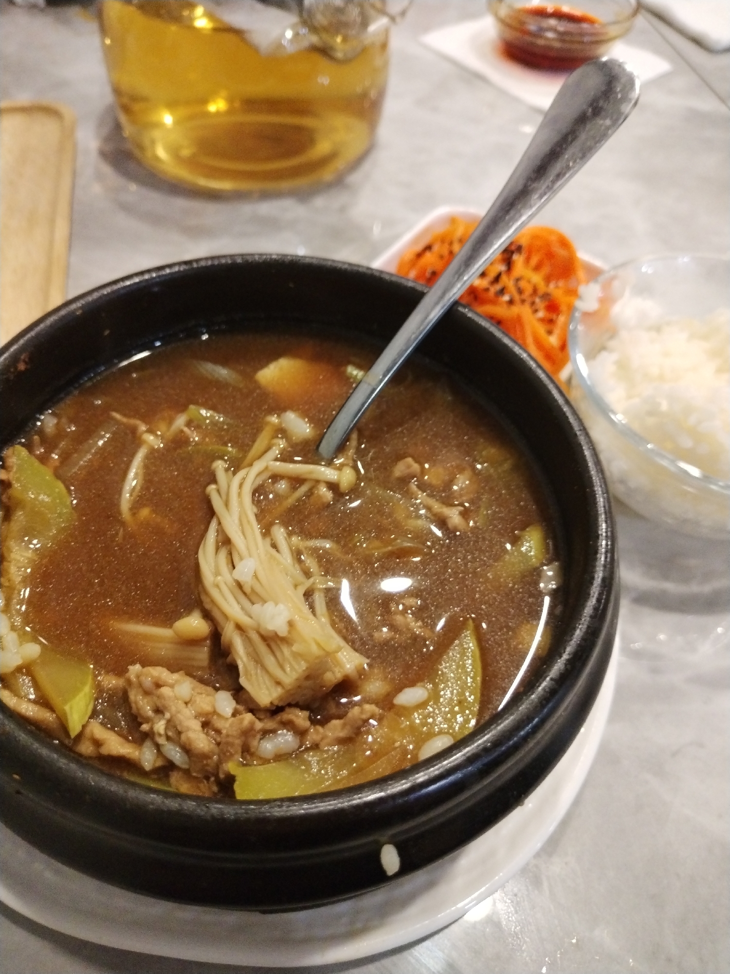 In Novonikol'sk, Russia, a close-up, top-down view captures a bowl of soup containing enoki mushrooms, thinly sliced meat, and green vegetables in a dark broth. A silver spoon rests in the soup. To the right, a small dish of shredded carrots and a bowl of white rice are visible. In the background, a glass pitcher filled with a golden liquid, possibly tea or beer, sits on a table with a speckled, light-colored surface. The scene appears to be set in a restaurant or dining area.