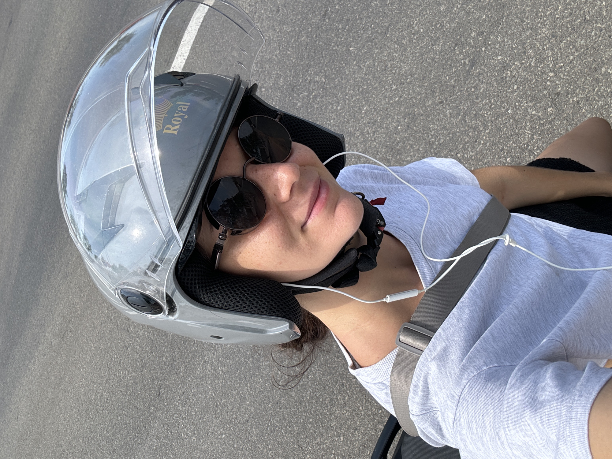 A medium shot shows a person wearing a gray helmet with a clear visor, round sunglasses, and a gray t-shirt, with a safety harness diagonally across their chest. The helmet has the word "Royal" on it. White wired earphones are visible. The background shows gray asphalt. The location is consistent with an urban area in Mui Ne, Vietnam, based on the visual style of the road surface and the nature of the transport safety gear.