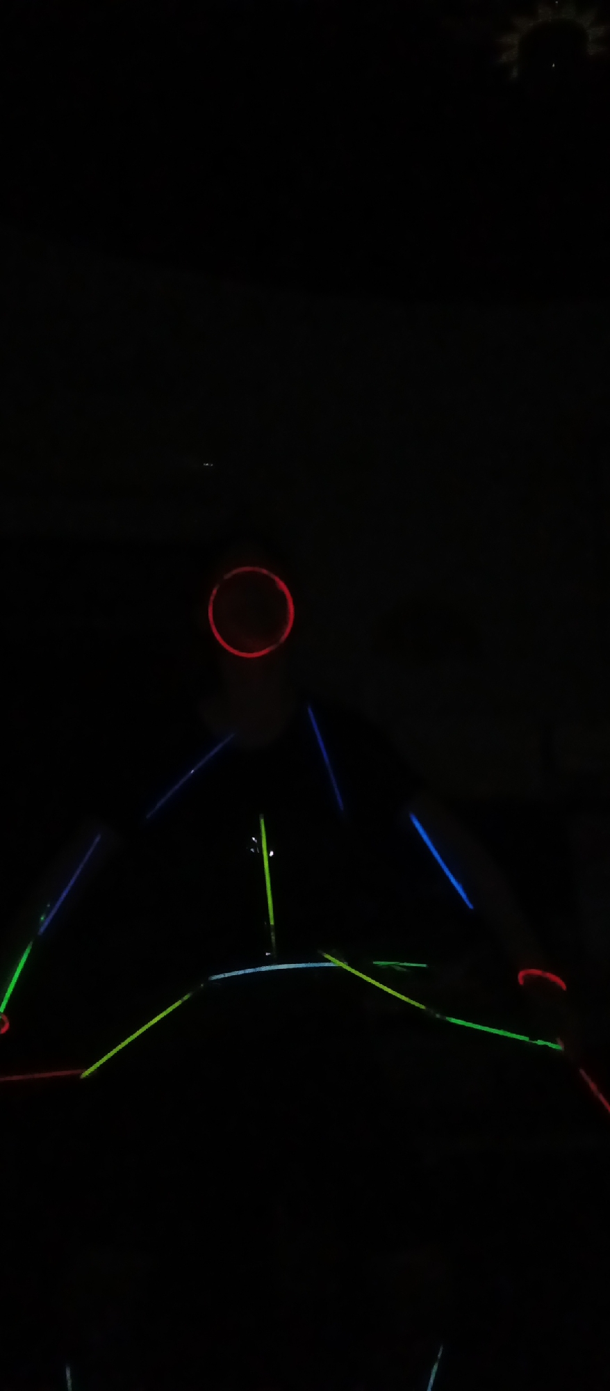 The image depicts a dark, indoor environment dominated by glowing light elements. At the top center, a prominent red glowing circle serves as a focal point. Below this, numerous glowing sticks or tubes in vibrant green and blue create a geometric structure, appearing to form a triangular or pyramidal shape. These luminous lines extend outwards and downwards, creating interconnected patterns across the frame.
The overall darkness suggests a low-light setting, possibly an indoor game or attraction such as a laser maze or a glow-in-the-dark activity. The glowing lines likely represent barriers or paths within this interactive space. Additional glowing red, curved shapes are visible towards the lower left and right edges of the image, potentially indicating other interactive elements or boundaries.
In the upper right corner, a faint, intricate silhouette resembling a sunburst or cog is barely visible against the dark ceiling, suggesting an overhead fixture. The scene is devoid of people, emphasizing the abstract and luminous nature of the installation. No discernible text is visible. The composition focuses on the contrasting bright, neon colors against the pitch-black background, creating an immersive and mysterious atmosphere.