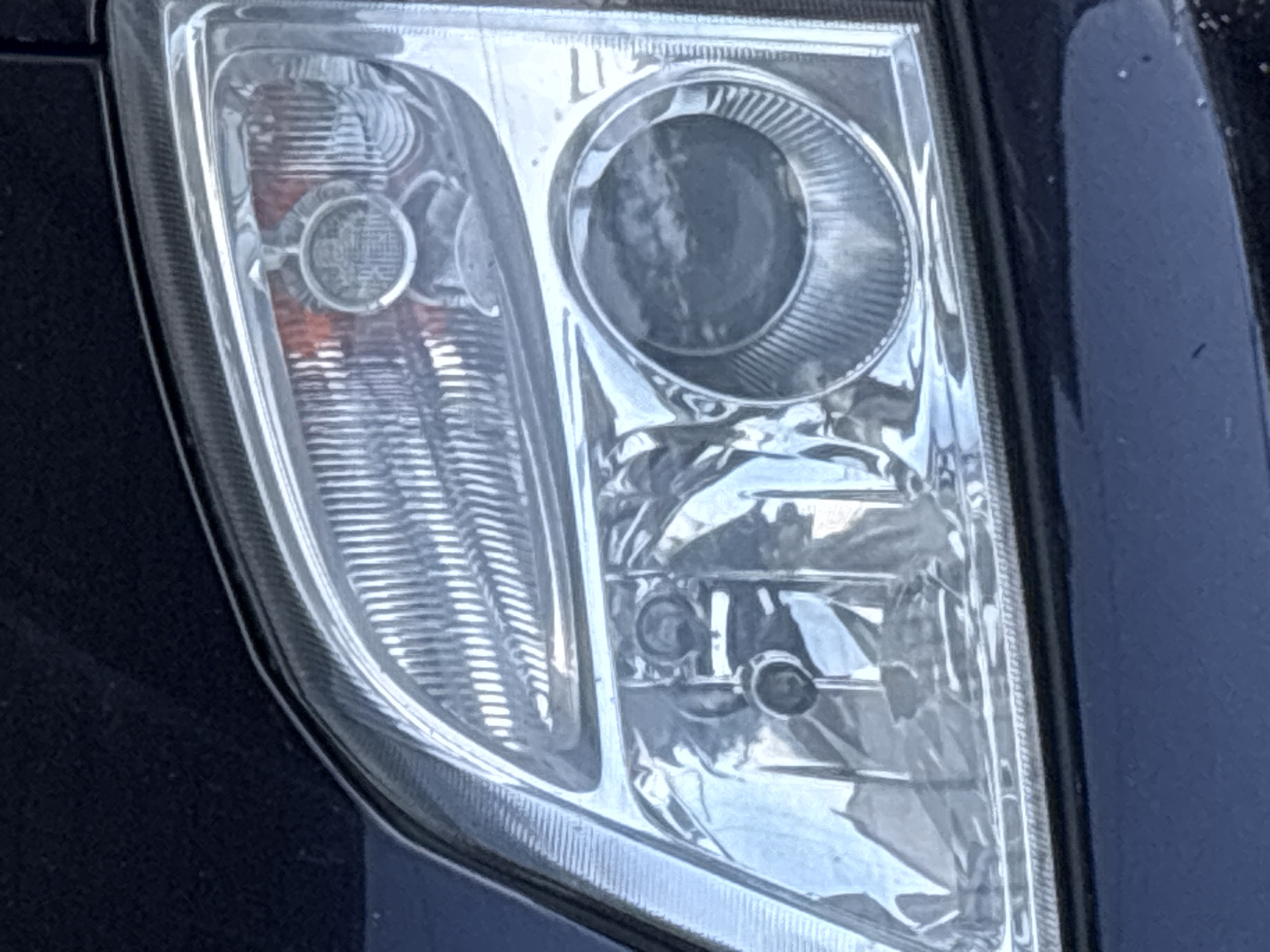 The image shows a close-up view of a car's headlight. The headlight is made of clear plastic with a ribbed interior and has multiple light bulbs visible. A dark blue car body is partially visible around the headlight. There is no indication of the city of Gurkovo, Bulgaria, or any people or actions in the image.