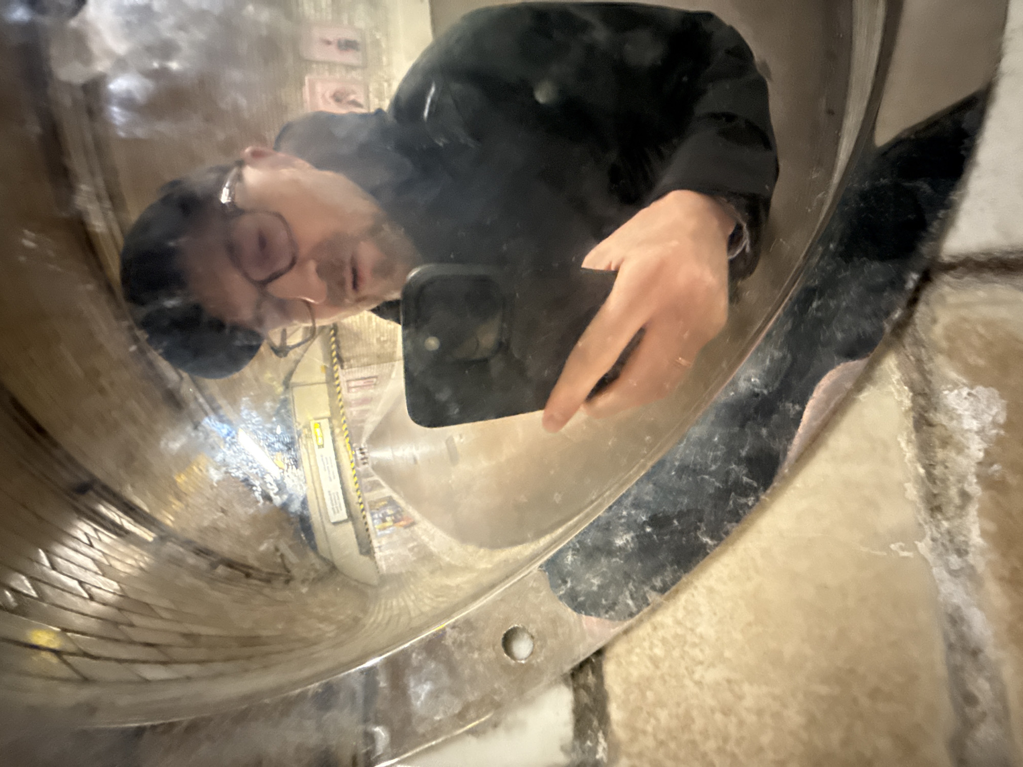 A mirrored self-portrait of a person wearing glasses and a black hat/beanie and black jacket is captured with a smartphone. The reflection reveals a cylindrical, tile-lined structure. The person is holding the phone to take the picture. The surroundings appear to be within the City Hall's "test tube" architectural structure in London, United Kingdom. Security/warning signs are visible in the background.