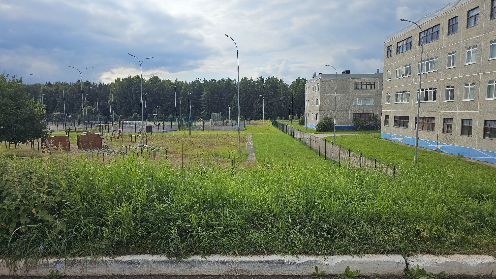 The image depicts a scene in Markovskii, Russia, featuring an outdoor recreational area adjacent to a multi-story apartment building. In the foreground, tall, lush green grass and weeds grow along a concrete curb. Beyond the grass, a well-maintained courtyard area is visible. This area includes a sports court, possibly for basketball, with visible hoops and boundary lines. Scattered throughout this area are various outdoor exercise equipment, including wooden structures resembling obstacles and metal bars arranged in a grid.
Further into the background, a dense line of trees forms a natural backdrop to the scene. Several tall lampposts are strategically placed throughout the courtyard, indicating it is likely lit for evening use. The apartment building, a light-colored, multi-story structure with numerous windows, stands prominently to the right of the image, its facade suggesting a functional, perhaps older, residential design. A black chain-link fence delineates the boundary between the courtyard and the grassy area. The sky above is overcast with dramatic clouds, suggesting a temperate climate. No people are clearly visible in the image, implying the area is either empty or the individuals present are too small or distant to be identifiable.