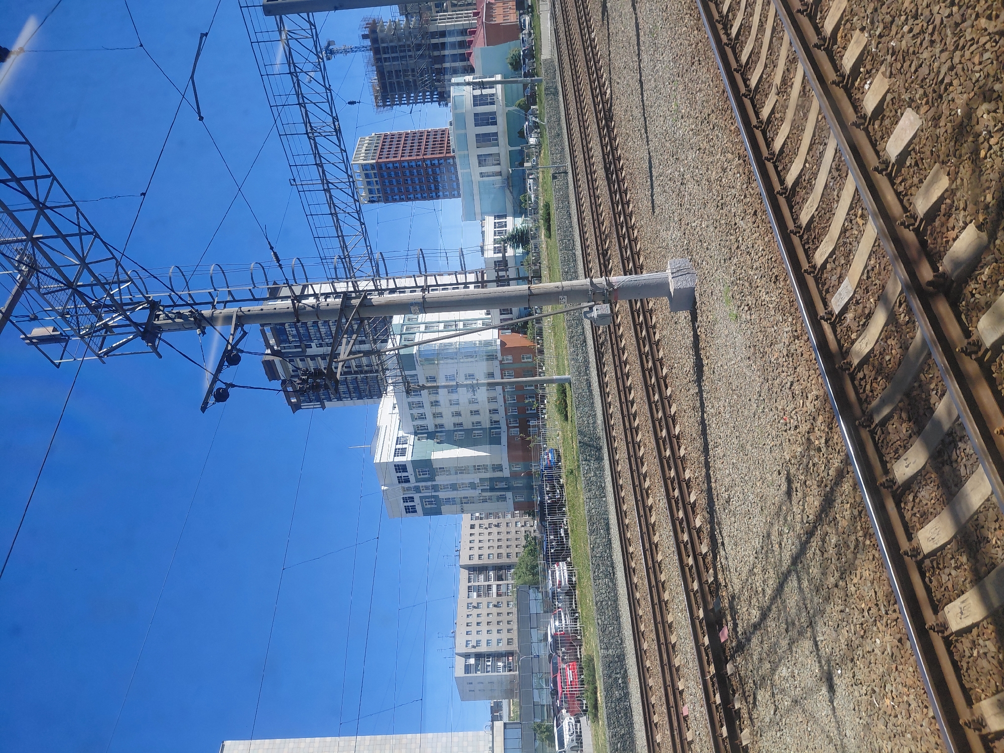 The image depicts a view of a city in Novosibirsk, Russia, seen from a train. In the foreground are multiple railway tracks, characterized by gravel ballast and concrete sleepers. Overhead, complex electrical catenary systems for trains are visible against a clear blue sky. Beyond the tracks, several modern buildings of varying heights and architectural styles are present. These include a tall, multi-story residential building with a white and light blue facade, another longer residential building with a beige facade and numerous balconies, and a taller, slender red-brown skyscraper with a grid-like pattern of windows. To the right, construction is underway on at least two high-rise buildings, indicated by cranes and unfinished structures. A parking lot with several cars is situated between the railway area and the buildings, separated by a fence. The overall environment appears to be an urban area with active infrastructure and ongoing development.