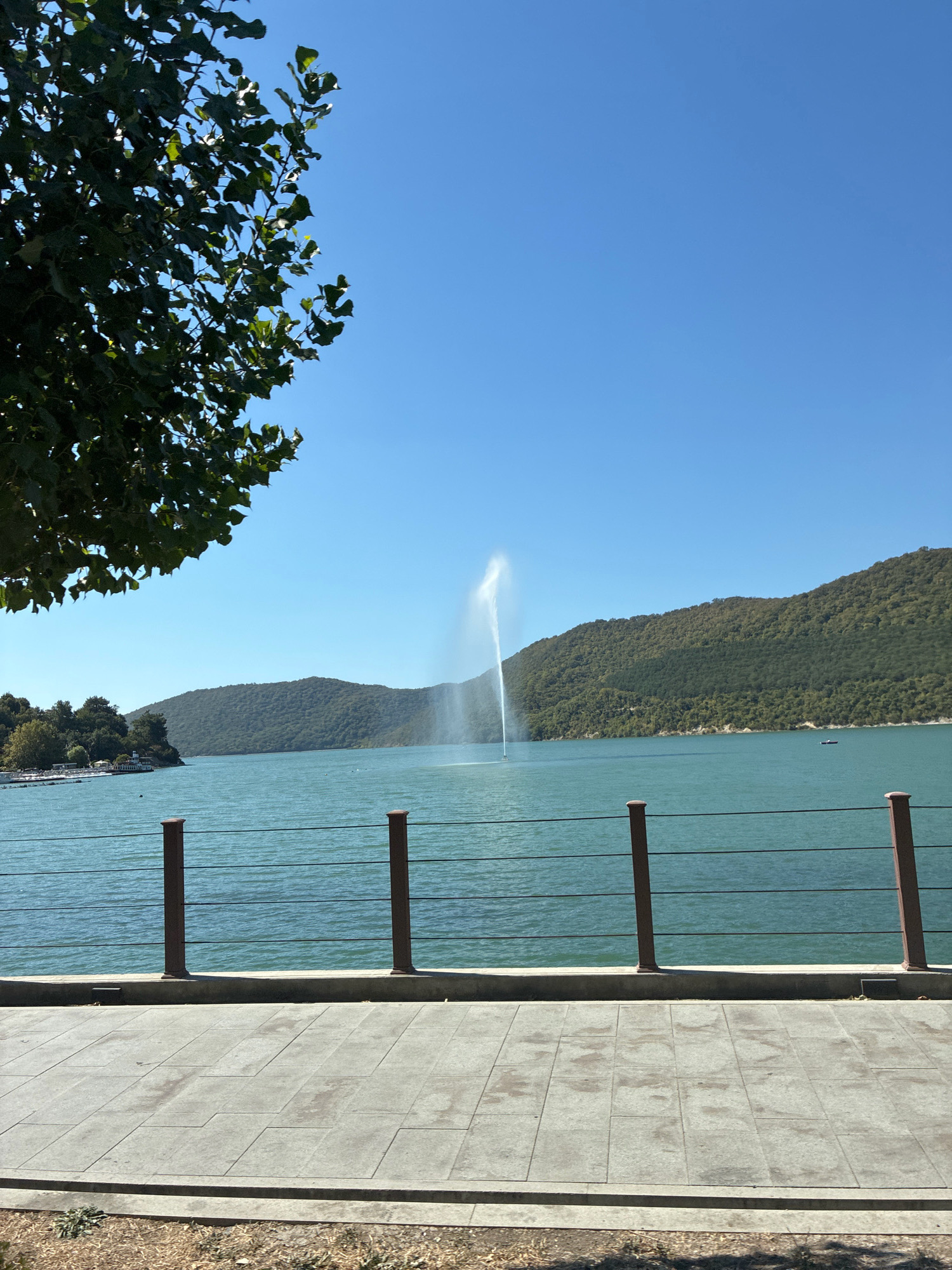 The image depicts a waterfront scene in Abrau-Dyurso, Russia, with a focus on Abrau Lake. In the foreground, a paved walkway runs parallel to a decorative railing, featuring metal posts and dark horizontal wires. Beyond the railing, the turquoise waters of Abrau Lake are visible, disturbed by a tall, arcing water fountain in the middle ground. The lake is bordered by lush, tree-covered hills that dominate the background, creating a picturesque natural setting. A clear, bright blue sky stretches across the upper portion of the image. On the left side of the lake, a shoreline with buildings and trees suggests the presence of a town or resort area. A small, dark boat is visible on the water to the right of the fountain, and another dark object, possibly a small boat or buoy, is near the left shore. A branch of a leafy tree partially obscures the upper left corner of the scene. The overall impression is one of a serene and scenic day at the lake.
