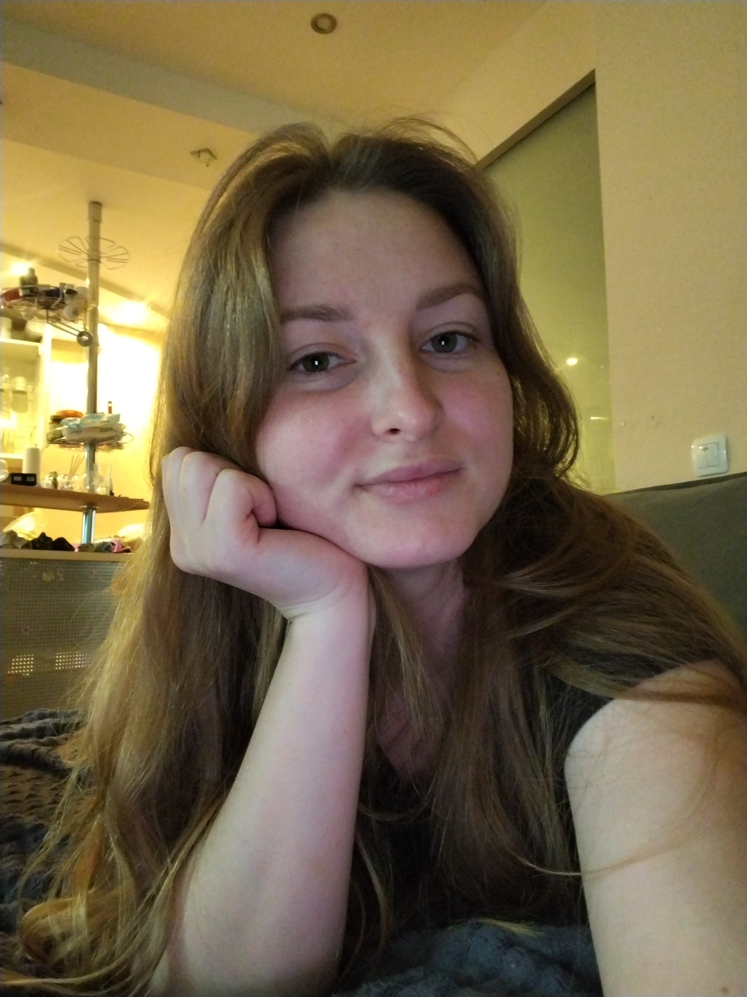 A woman with long, light brown hair is resting her face on her hand, likely sitting on a couch or bed. She is looking directly at the camera with a slight smile. The background includes a kitchen or dining area with a shelving unit containing various items and a hanging light fixture. A wall with a light switch is also visible. The image was taken in an indoor location, presumably a home or apartment. The image does not contain any information about the city of Chaikovsky, Russia.