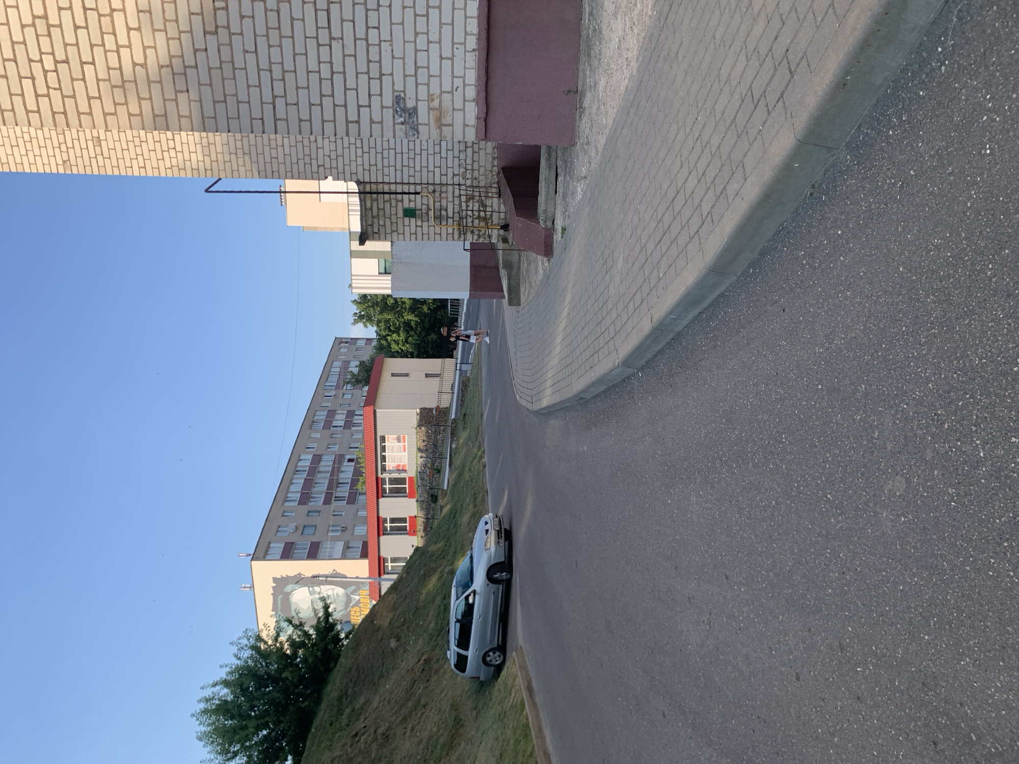 This image depicts an outdoor scene with apartment buildings and a street in Kapyĺ, Belarus. The main subjects are two apartment buildings, a silver car parked on the side of the road, and a pedestrian sidewalk made of brick pavers. The buildings have a mix of beige and gray facades, with windows of varying sizes. One building features a large mural of a person's face. The sky is clear and blue, suggesting it is daytime, likely late afternoon or early evening given the long shadows cast by the buildings and the warm lighting. The street is paved with asphalt, and a grassy slope is visible beside the road.
The scene appears to be a typical urban or suburban environment. The presence of apartment buildings suggests a residential area. The car is parked parallel to the curb, and there is a person in the distance walking on the sidewalk, indicating normal daily activity. No specific event is taking place, but the setting implies a peaceful, everyday moment. The lighting and clear sky create a sense of pleasant weather.
There is some visible text on the side of the building with the mural, appearing to be in Cyrillic, though it is not entirely legible. The overall impression is one of a quiet, ordinary day in a residential area of Belarus.