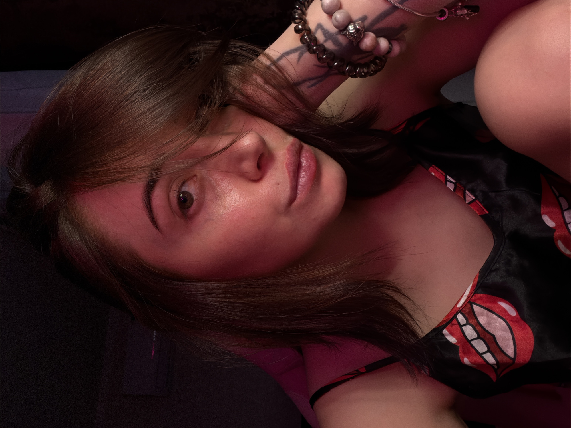 A young woman is captured in a close-up, head-and-shoulders shot, bathed in soft, ambient pink light. She appears to be indoors, in what seems to be a bedroom, with a dark background and a hint of an air conditioning unit visible on the wall. The woman has medium-length, brown hair that partially obscures her face, with strands falling across her forehead and cheek. Her eyes are open, looking directly at the camera. Her lips are slightly parted. She is wearing a black camisole with a bold, red and white lips print. On her left wrist, she wears several beaded bracelets, and a tattoo is visible on her forearm. Her left arm is resting near her face, supporting her head, and her right leg is bent, with her knee close to her chest. The overall impression is one of casual intimacy, with the lighting creating a somewhat moody and artistic atmosphere.
