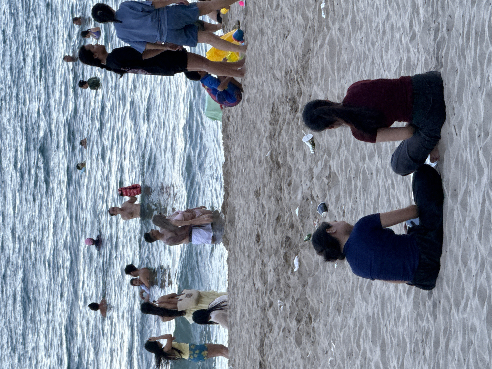 The image depicts a crowded beach in Nha Trang Ward, Vietnam, with numerous people enjoying the water and the sand. In the foreground, two women are seated on the sand, facing away from the camera. They are both wearing casual clothing. The beach itself appears somewhat littered with discarded cups and debris.
Further into the scene, the ocean is filled with people swimming and wading. Several individuals are visible in the water, some appearing to be playing or socializing. Various colored buoys and inflatables are scattered throughout the water.
On the shoreline, a group of people are standing in the water, some with children. A person in a black t-shirt and shorts is standing near the water's edge, facing towards the sea. Another individual in a blue t-shirt and shorts is also visible. Near them, a young child in a blue and red swimsuit is crouching on the sand.
The water is a grayish-blue, indicating potentially overcast weather or the time of day. The sand is a light brown, with ripples and patterns created by the wind or waves. The overall impression is a lively beach scene, though the presence of litter detracts from the natural beauty of the environment.