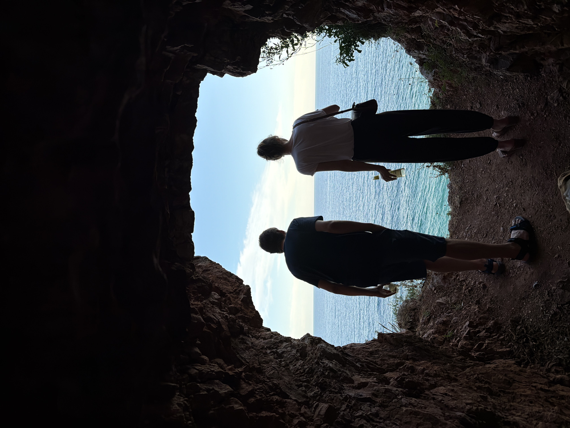 Two people, a man and a woman, stand silhouetted within the dark entrance of a cave, looking out at the bright expanse of the Adriatic Sea. The man, on the left, wears a dark t-shirt and shorts, with sandals. He holds a clear bottle in his left hand. The woman, on the right, wears a white t-shirt and dark pants, with sandals. She carries a dark bag over her shoulder and holds a clear bottle in her right hand. Their figures are outlined against the luminous scene of the sea and sky, with the cave's rough, textured rock walls framing the view. The water is a vibrant turquoise, meeting a pale blue sky with scattered clouds. The ground within the cave appears to be dirt and small stones, with some sparse vegetation visible at the edges. The scene is one of quiet observation, with the two individuals contemplating the seascape from their sheltered vantage point. This location is likely a sea cave near Petrovac centar, Montenegro.
