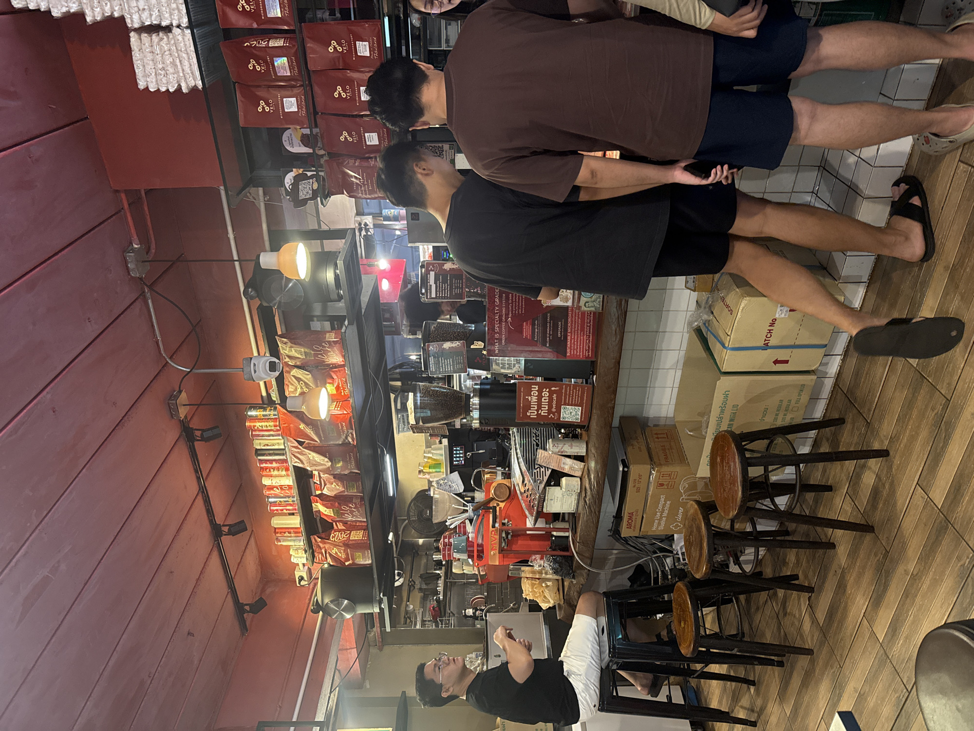 The image depicts the interior of a coffee shop, likely located in Hua Hin City Municipality, Thailand, based on the Thai text visible. The scene is viewed from a medium distance, capturing a counter area with people and coffee-related equipment.
In the foreground, several wooden stools with metal legs are arranged on a wooden floor. To the left, a man wearing a black t-shirt and white shorts is seated on a black stool, looking towards the counter. He appears to be wearing glasses.
Behind the counter, a young man in a black t-shirt and dark shorts is standing with his back to the camera, facing the counter. Another young man, wearing a brown t-shirt and dark shorts, stands next to him, also facing the counter. Further to the right, a person's arm and shoulder are visible.
The counter itself is made of wood and displays various items, including coffee bags, a coffee grinder, and what appears to be a coffee machine. There are shelves above the counter stocked with bags of coffee beans, some with visible branding and Thai text. Various boxes and other items are stacked on the floor behind the counter. The overall lighting suggests an indoor environment, with overhead lights and a warm ambiance. The walls and ceiling are painted in muted tones, with the ceiling featuring a wooden plank design.