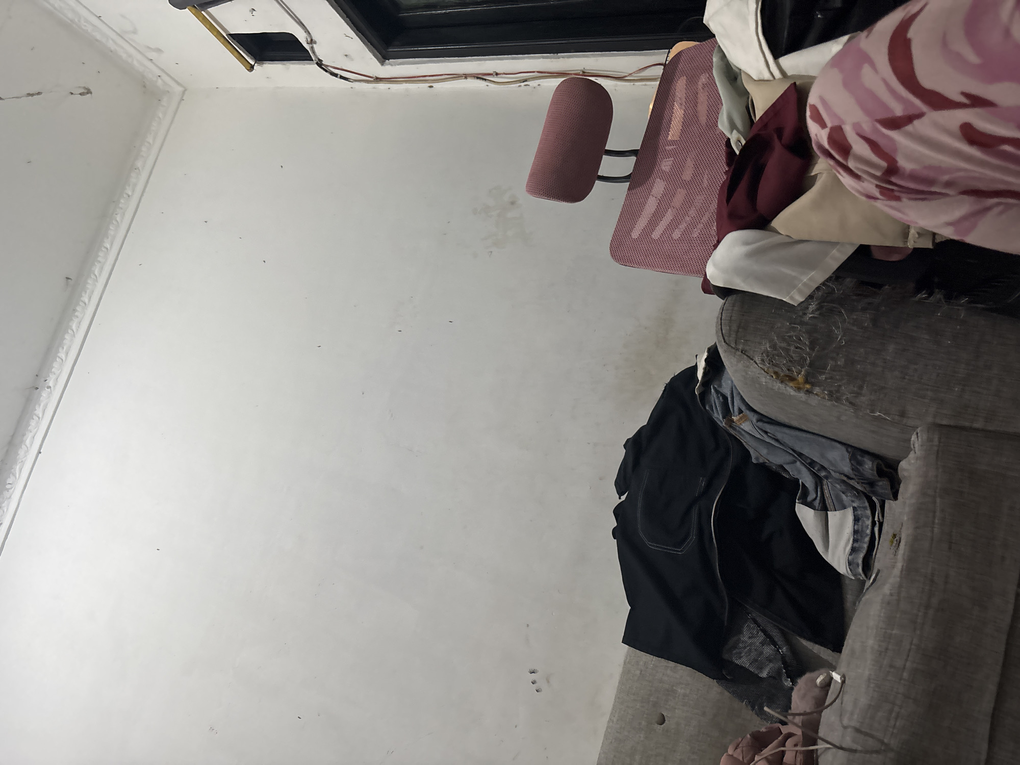 The image depicts a messy interior scene, likely an apartment, located in North Jakarta, Indonesia. In the foreground, a grey sofa is covered with various articles of clothing, including a black jacket, denim jeans, and other folded garments. The sofa appears worn, with some visible damage to its fabric. To the right of the sofa, a pink mesh office chair with a headrest is positioned. The walls are white and show some signs of wear and tear, with visible marks and a decorative molding along the top. A doorway with black trim is visible on the far right, with electrical wiring running down the wall beside it. The lighting suggests an indoor, possibly dimly lit environment.