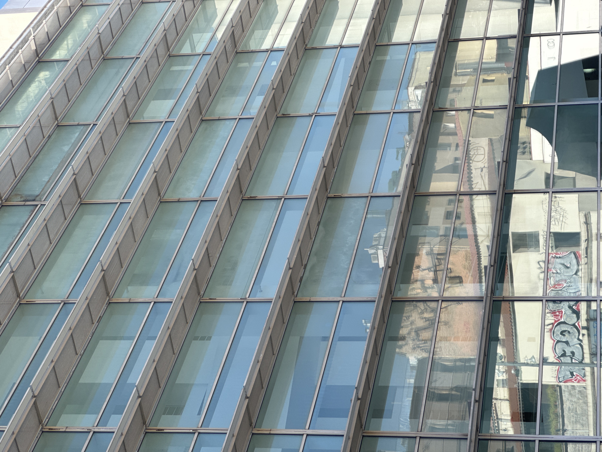 The image shows a section of the glazed facade of the "Torre Glòries" in Barcelona, Spain. The diagonal arrangement of the windows creates a pattern. The windows on the right reflect the urban landscape, including a building with graffiti.