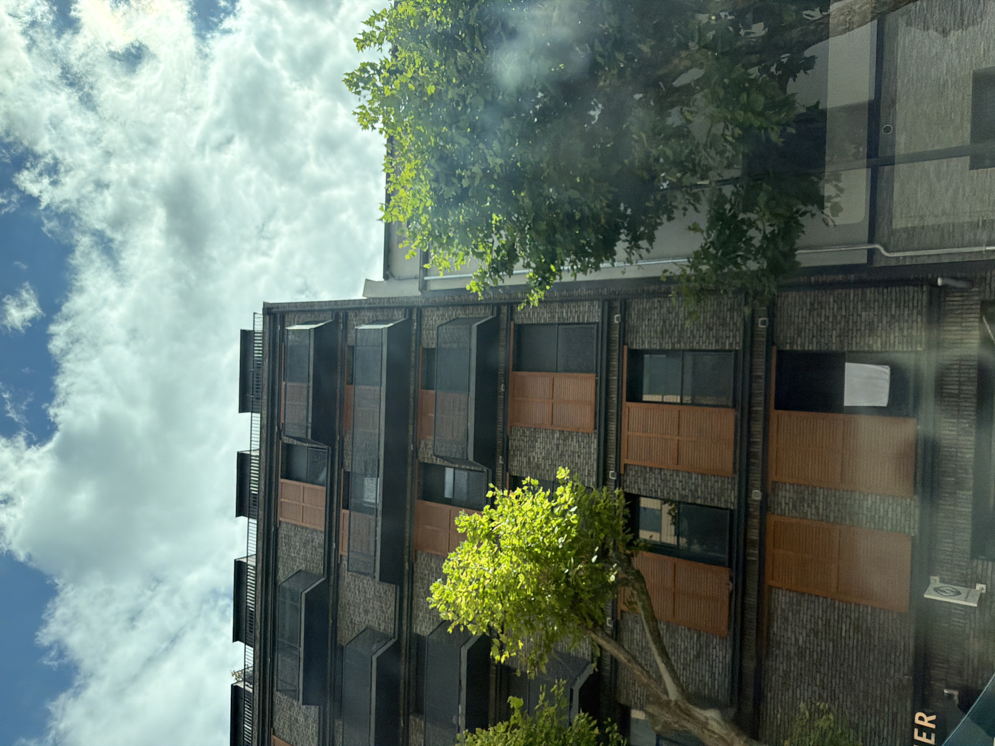 This is a medium shot, eye-level, exterior photograph of a modern brick building with multiple balconies. The building is tall, with several stories visible. The balconies are dark colored with glass railings. Many windows have wooden shutters that are a reddish-brown color and appear to be closed. Two trees with bright green leaves are in the foreground, one on the left and one on the right side of the frame. The sky above is blue with fluffy white clouds. A white towel or sheet is hanging from a balcony on the right side of the building. The bottom of the image shows the facade of the lower part of the building, including what appears to be a sign that reads "ER" in white capital letters on a dark background, and other architectural details. The image appears to have been taken from a street level perspective, possibly from a vehicle, given the slight glare on the left side of the image. The architectural style suggests a contemporary urban setting.