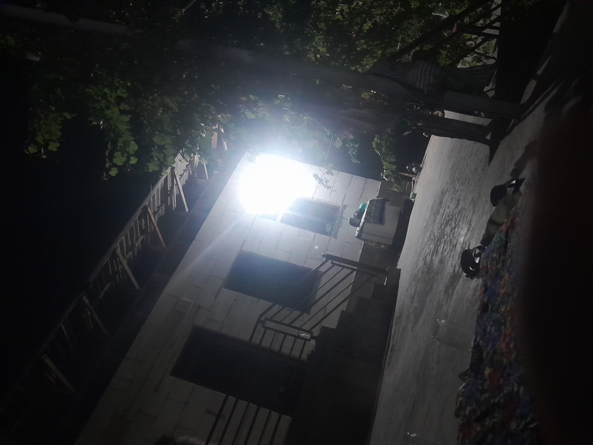 A nighttime exterior shot of a building under construction in Chartak, Uzbekistan, is shown. The building's exterior walls appear to be constructed of white cinder blocks, with exposed wooden framing for the roof. A set of stairs with a metal railing leads up to a doorway. A bright light emanates from a window, illuminating the area in front of the building. To the right of the building, a tree with dense foliage is visible. The ground in front of the building is paved. In the foreground, a colorful blanket or rug lies on the ground, with what appear to be two shoes or slippers on top of it. A glass jar is also visible on the paved surface. The scene suggests a residential area in Chartak, Uzbekistan, possibly in a rural or suburban setting given the uncompleted construction and the presence of a large tree. The lighting indicates it is nighttime, with artificial light sources illuminating the scene.