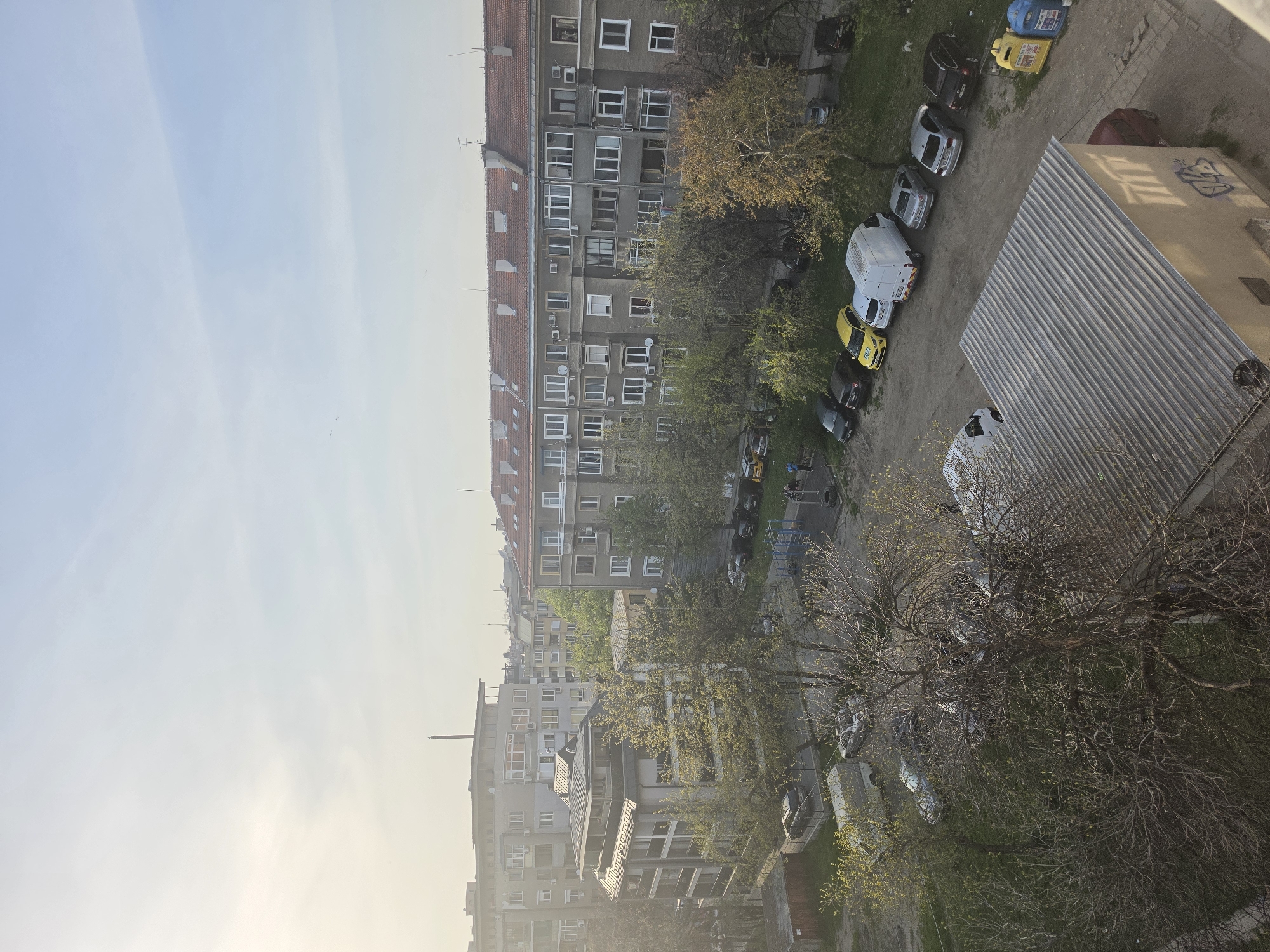 The image shows an aerial view of a residential area in Plovdiv, Bulgaria. Several apartment buildings are visible, with a tall building with a red roof dominating the view. Trees are interspersed between the buildings and along the street, some just beginning to show new leaves. Cars are parked along the side of the road. A large corrugated metal roof covers a structure in the foreground, casting a shadow. In the distance, the sky is pale blue. There are recycling bins near a parking area.
