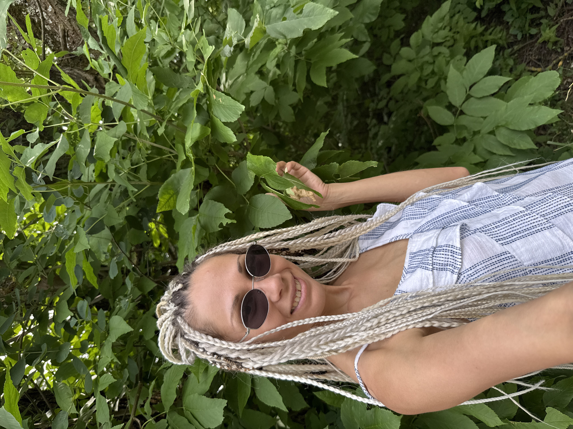 A young woman with blonde braided hair and sunglasses smiles at the camera while standing outdoors in a lush green natural environment. She is wearing a white and blue striped dress. Her right hand is raised and holding a cluster of green leaves. The background is filled with dense green foliage and the bark of a tree is visible on the right side of the image. The overall impression is one of a relaxed, outdoor portrait. The location is identified as Rusanovo, Russia.