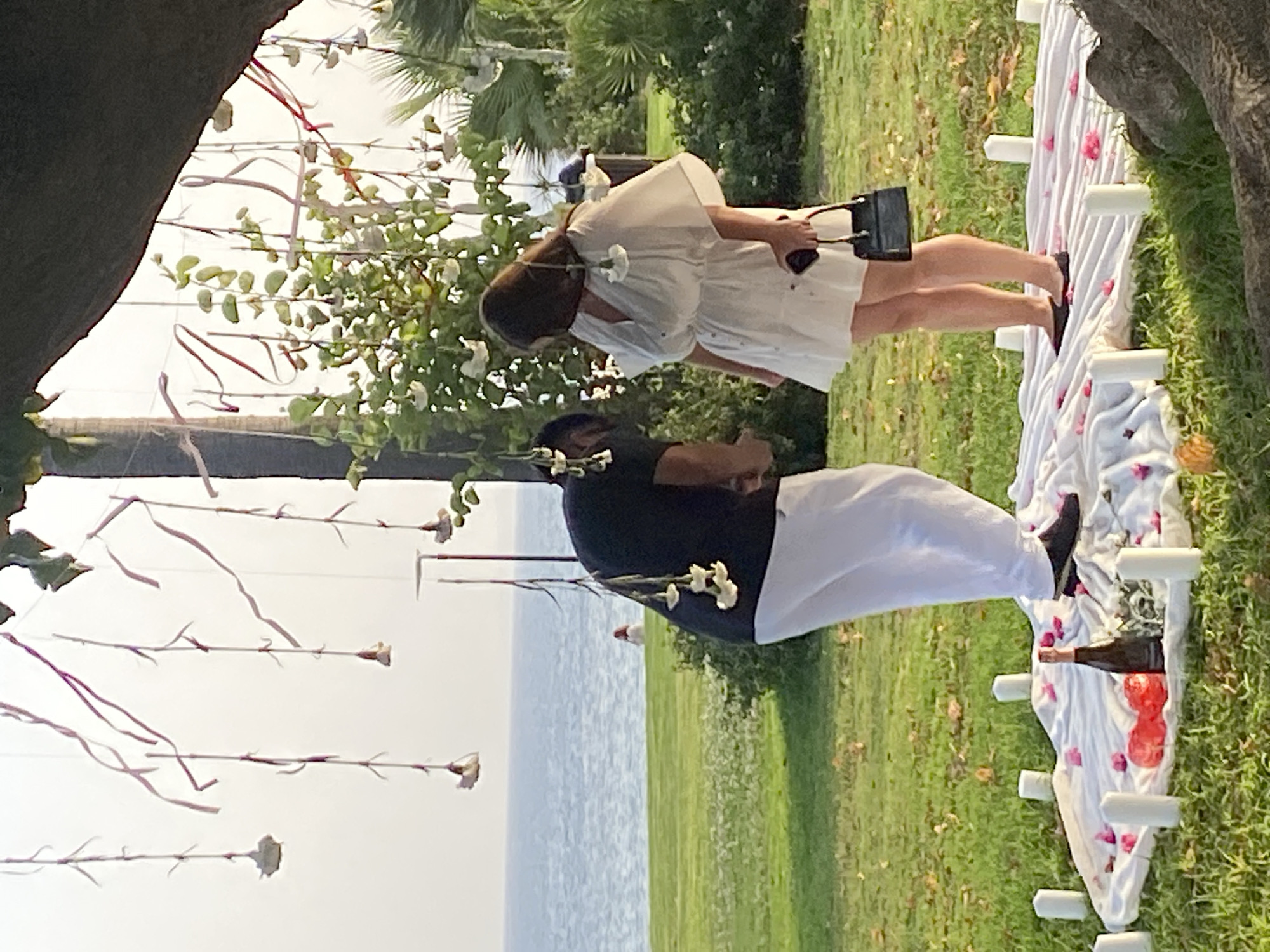 Two individuals, a man and a woman, are positioned outdoors in a grassy area overlooking the sea, possibly in Estepona, Spain, near a beach. The setting appears to be decorated for a romantic occasion. The man, wearing a black t-shirt and white pants, is leaning forward, facing the woman. The woman, in a white mini-dress and black flat shoes, stands facing him, holding a black purse. In front of them, on a white cloth spread on the grass, are several white pillar candles, two red decorative objects, and a bottle of wine, with scattered pink flower petals. Hanging from overhead branches are delicate decorations of small white flowers and pink ribbons. The background shows the blue sea under a bright sky, with green grass and palm trees.