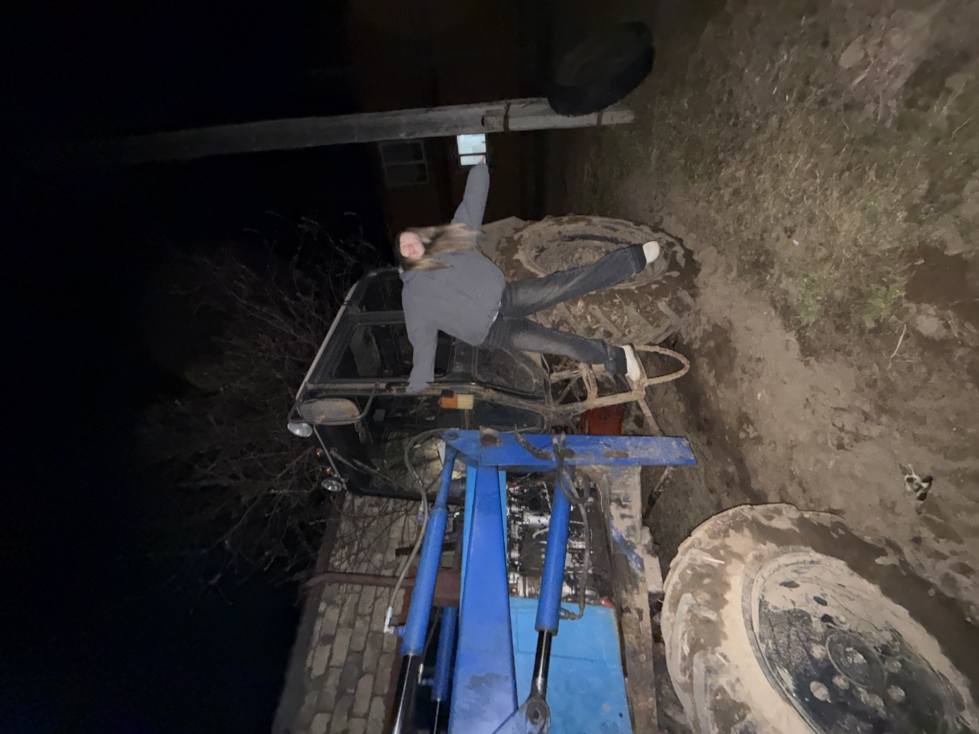 The image shows an individual lying on top of a tractor cab. The tractor is outdoors on a dirt and grassy area, with a jack apparatus connected to it. It is nighttime, and the lighting is poor. A building is visible in the background. Based on geolocation, it is possible to assume this is Zhigalki, Russia, although there is no geotag in the original data. The individual is wearing dark jeans, light-colored shoes, and a gray jacket. The person's left arm is raised.
