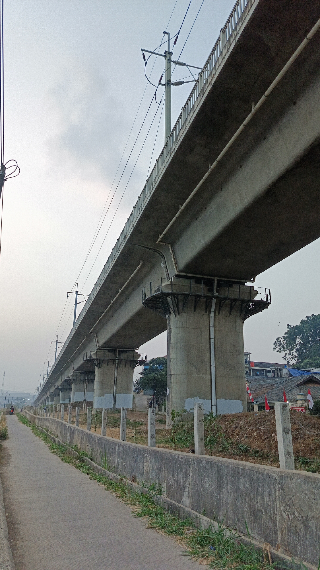 The image depicts a long, elevated concrete structure, likely a viaduct or elevated railway, stretching into the distance under a hazy, overcast sky. The structure is supported by numerous concrete pillars. A paved walkway runs alongside the structure, bordered by a low concrete barrier and sparse vegetation. In the background, buildings and trees are visible, suggesting an urban or semi-urban environment. A single individual on a motorcycle is visible on the walkway in the far distance. Indonesian flags are displayed on one of the buildings in the background. The scene appears to be located in Margajaya, Indonesia, as indicated by the context provided.